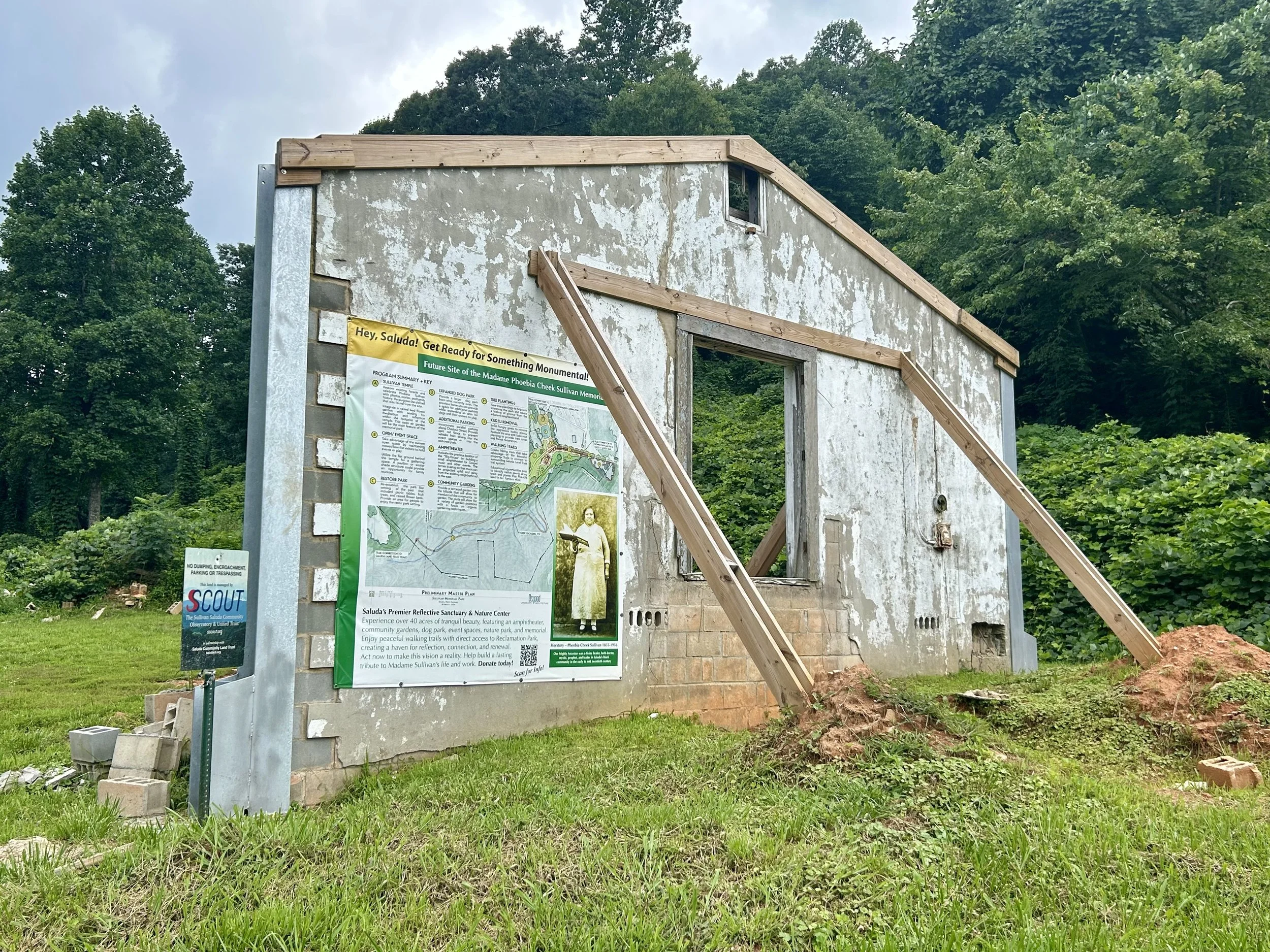 A partially constructed building with a sign and informational board in front, surrounded by trees and greenery.
