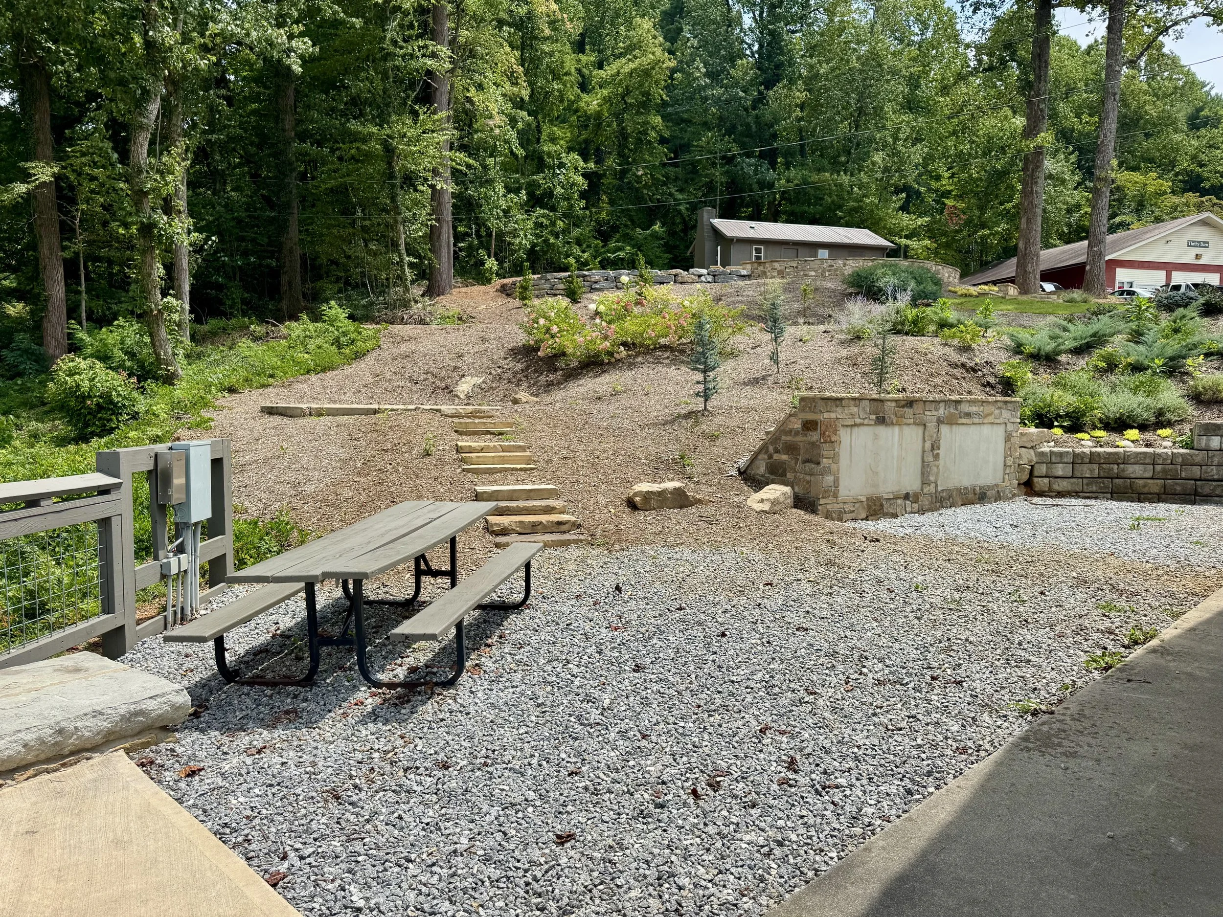 A backyard with gravel ground, a picnic table near a concrete path, and a stone retaining wall with plants. In the background, there are tall trees, a small gray shed, a house, and parked cars.