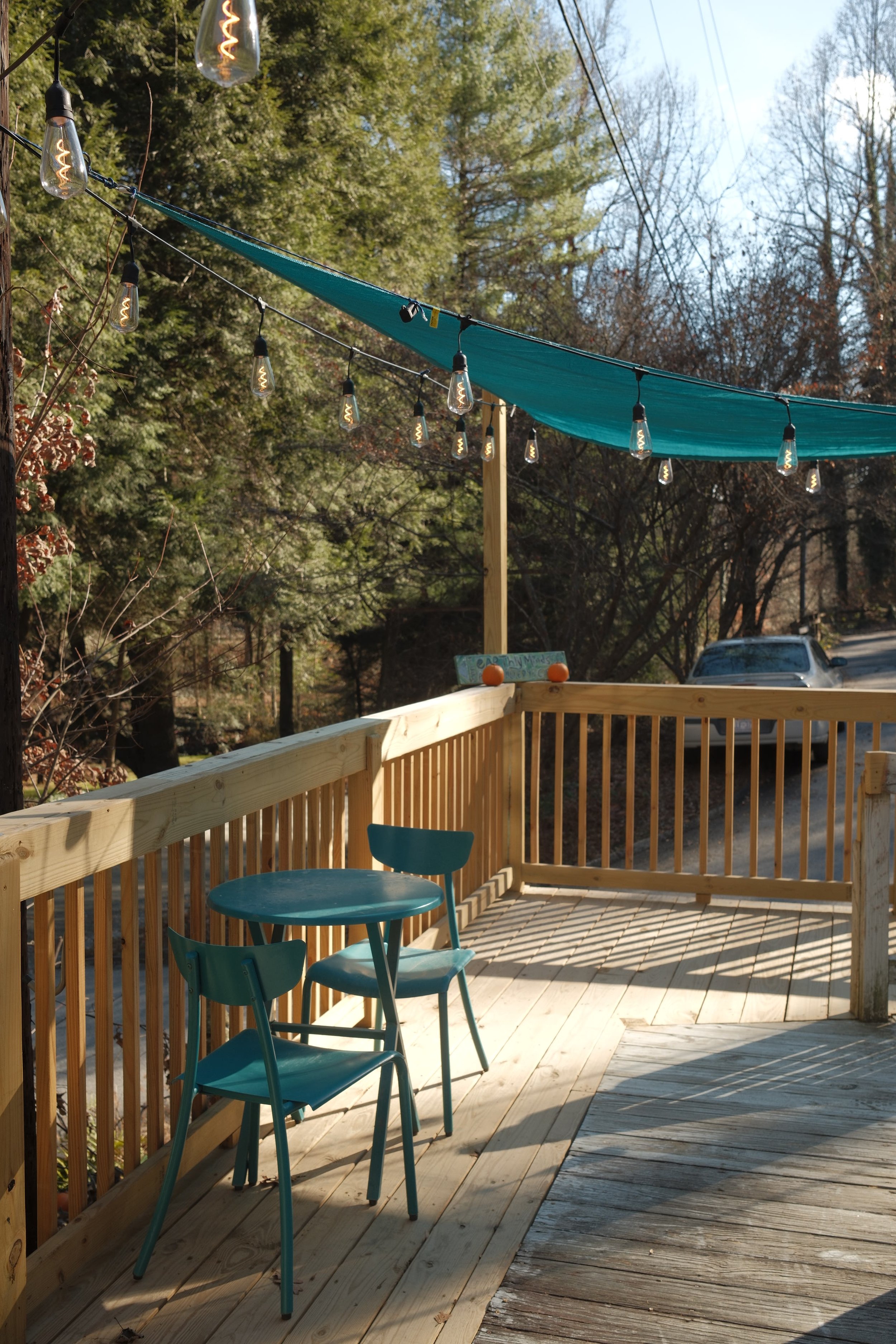 An outdoor patio with a wooden railing, blue chairs, and a round blue table. String lights hang above, and a teal shade cloth is stretched overhead. Trees and a car are visible in the background on a sunny day.