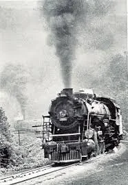 Black and white photo of a steam locomotive chugging along a railway track with smoke billowing from its chimney.