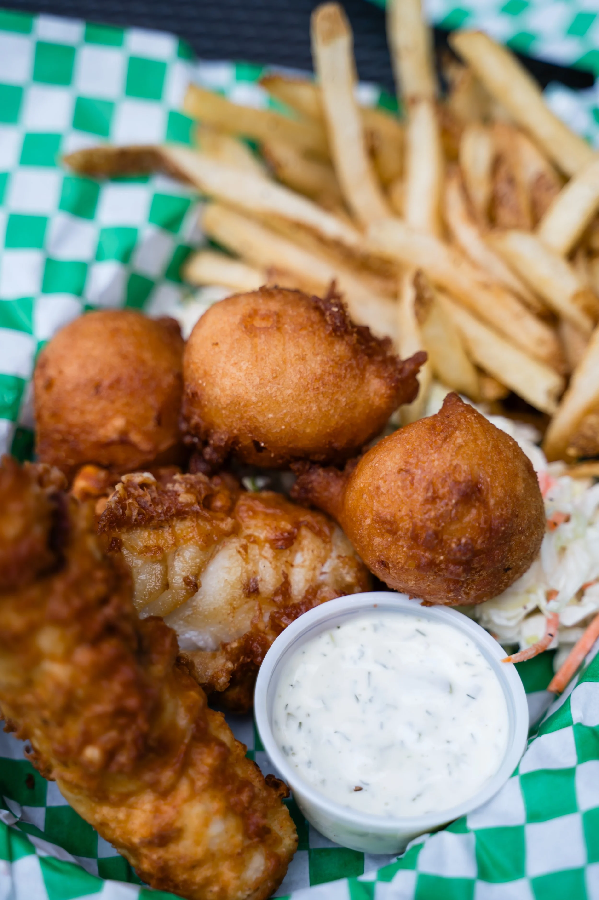 Fried fish fillet, hush puppies, French fries, coleslaw, and tartar sauce on green and white checkered paper.