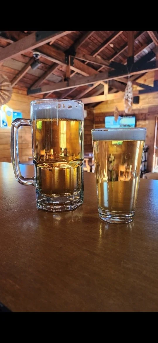 Two glasses filled with beer placed on a wooden table in a rustic bar or restaurant with wooden interior and TV screens in the background.