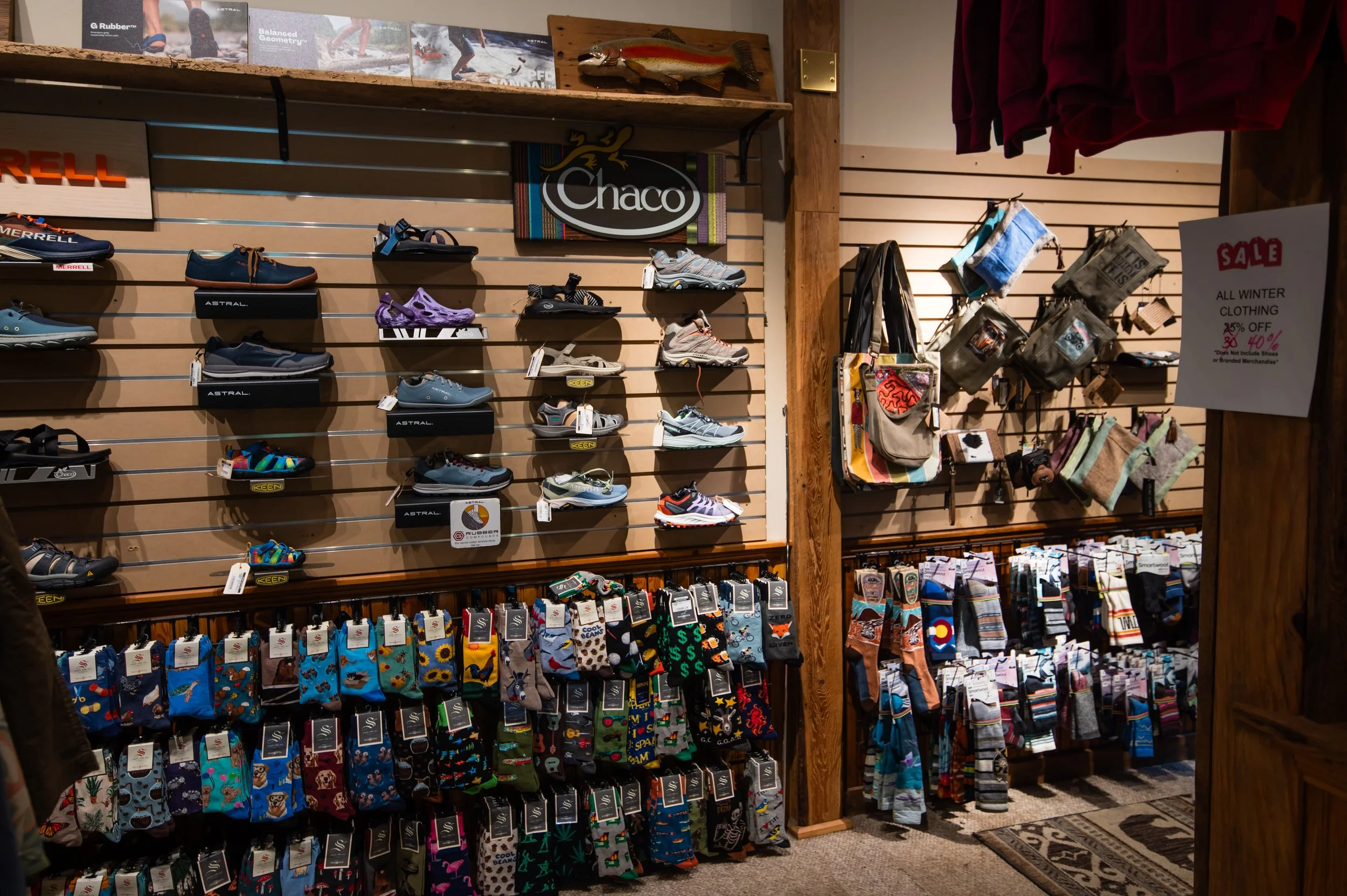 A retail display shelf with various shoes, including athletic and casual styles, arranged on slatwall panels. There are also hats, socks, and a sale sign with discounts on winter clothing and shoes.