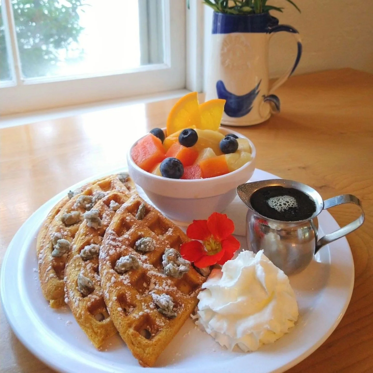 A breakfast plate with waffles topped with powdered sugar and nuts, a bowl of mixed fruit, a dollop of whipped cream, a small metal pitcher of syrup, and a cup of black coffee. There's a red flower garnish on the plate. In the background, a ceramic p
