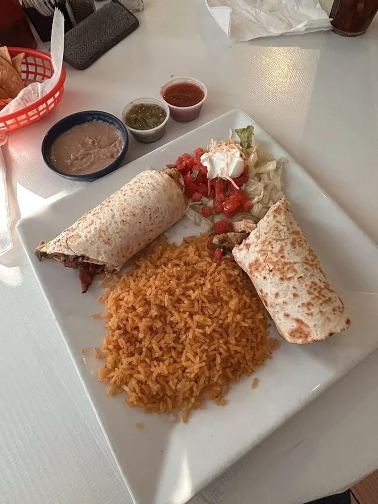 A plate of Mexican food with two burritos, Mexican rice, and a small salad topped with sour cream, along with three small bowls of salsa and guacamole.