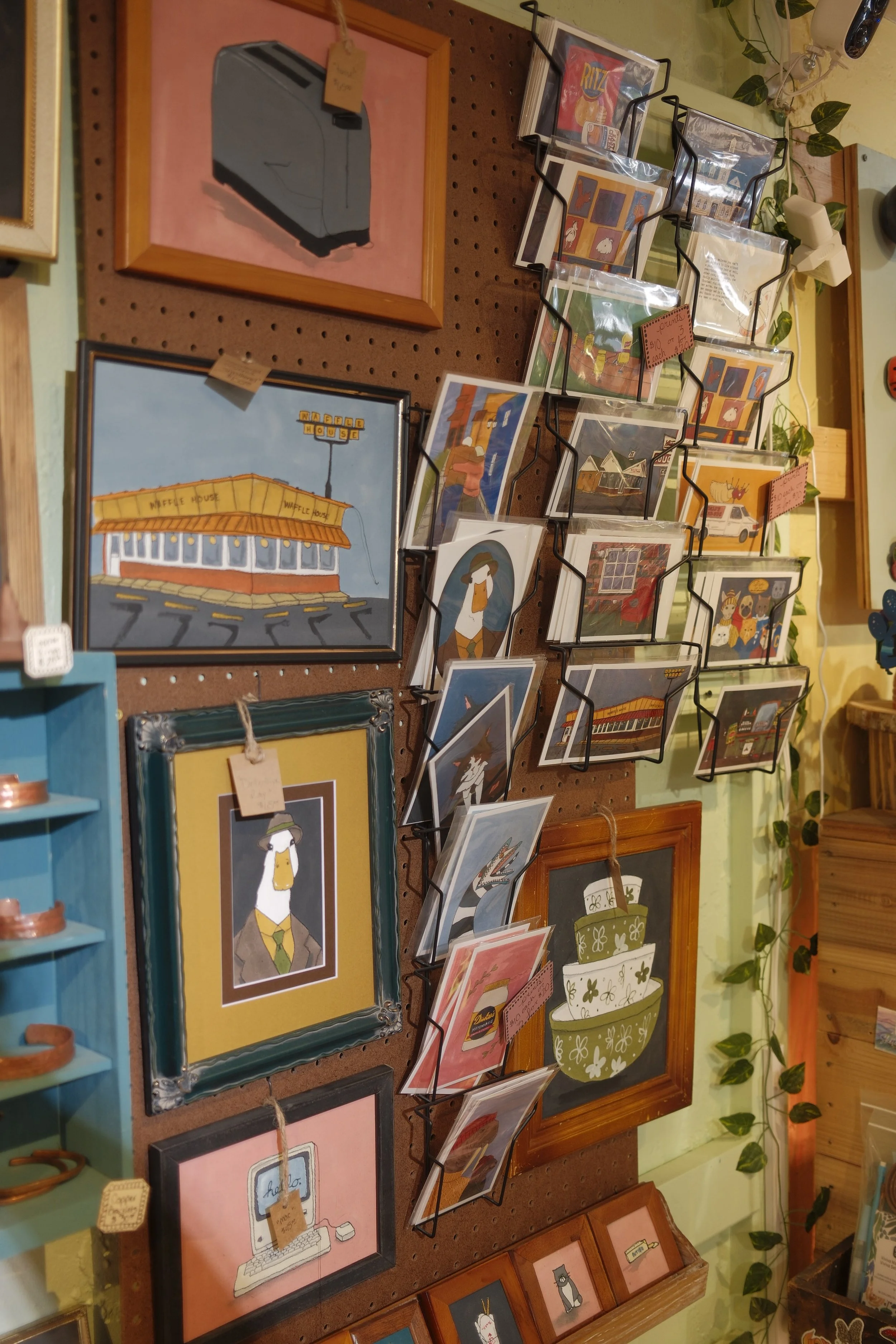 A display of framed illustrations on a pegboard, including a gray typewriter, a 'Waffle House' sign, and various artwork displayed in stands.