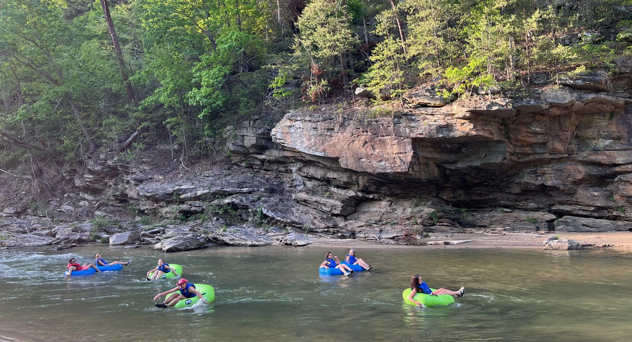 People floating on inflatable tubes in a river near a rocky shoreline with dense green trees and cliffs in the background.