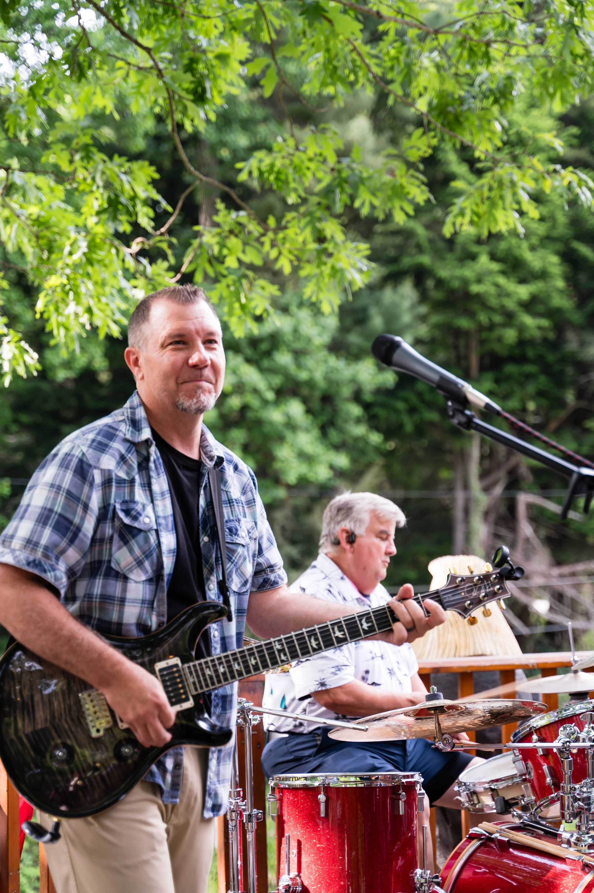Two men playing musical instruments outdoors under trees. One man is standing and playing an electric guitar, while the other is sitting and playing drums. The setting appears to be a casual outdoor gathering or performance.