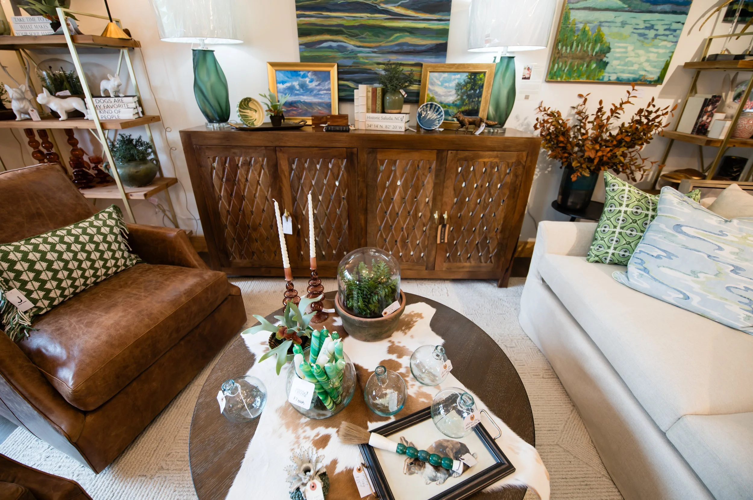 Living room with a brown leather couch, a white sofa with green and blue patterned pillows, a cowhide rug, and a wooden dresser decorated with paintings, vases, and books. There are shelves with decorative items including dog figurines, plants, and a