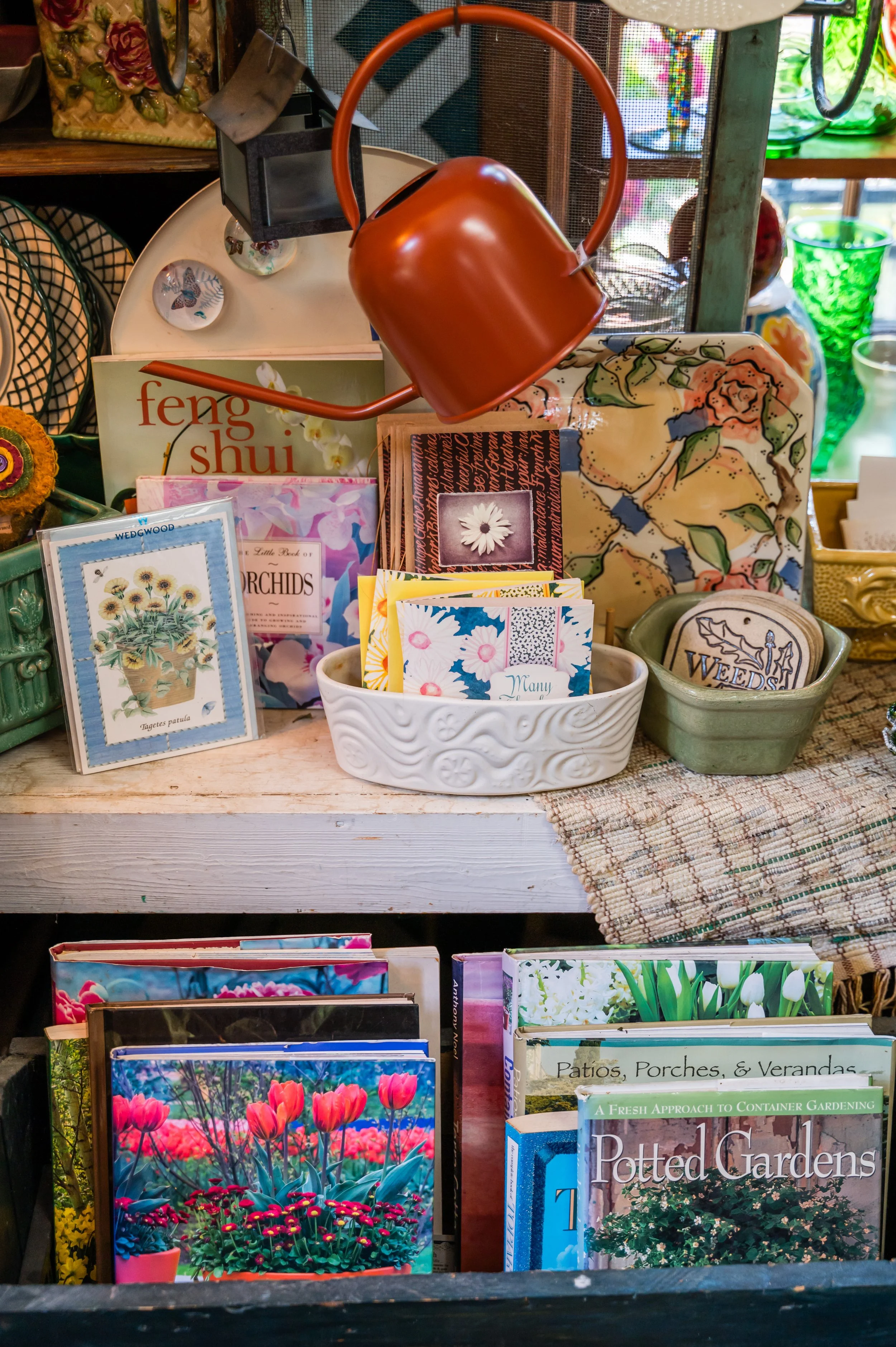 A display of gardening books and decorative items on a wooden shelf. There are colorful flower photographs, gardening guides, and ceramic containers, along with a rust-colored watering can and other garden-related items.