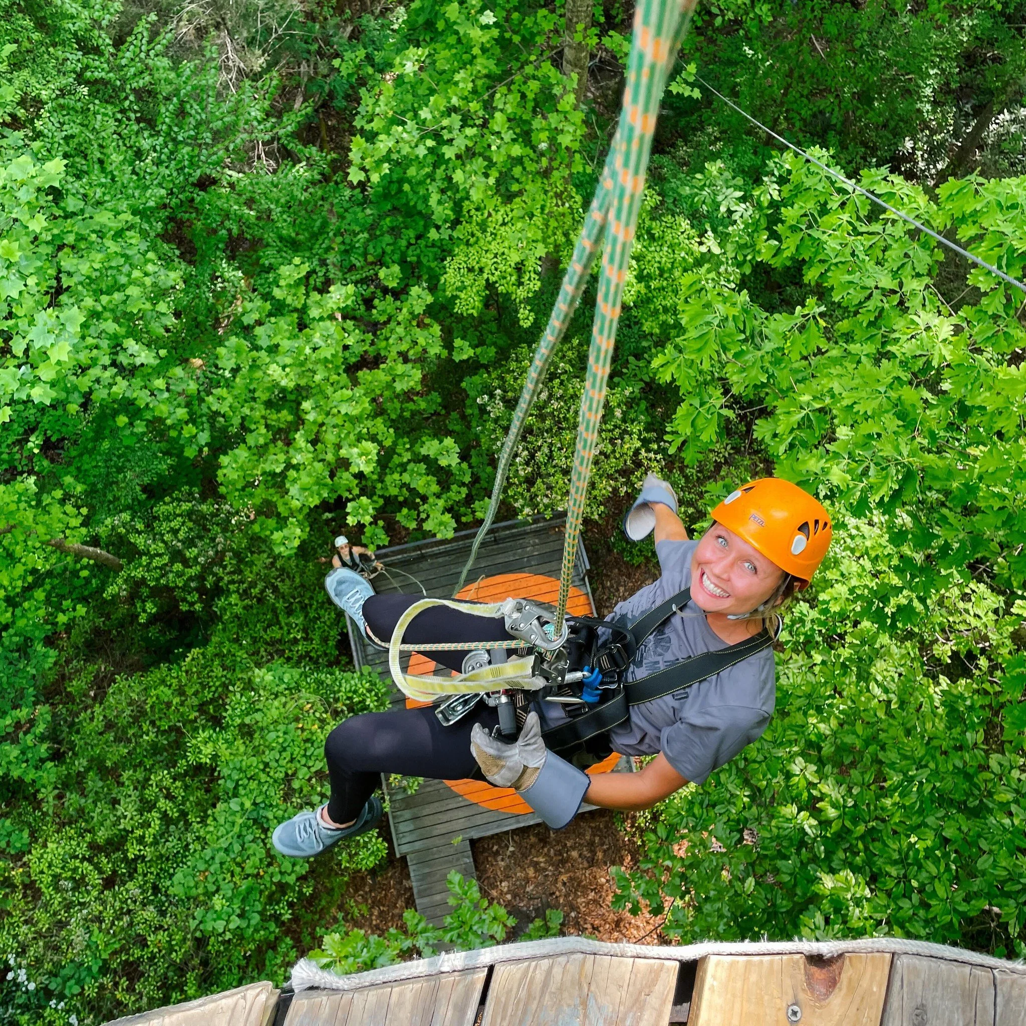 A woman wearing an orange helmet and gray shirt is smiling at the camera while sitting on a tree platform. She is balancing on a safety harness connected to a rope, surrounded by dense green leaves and branches in a forested area.