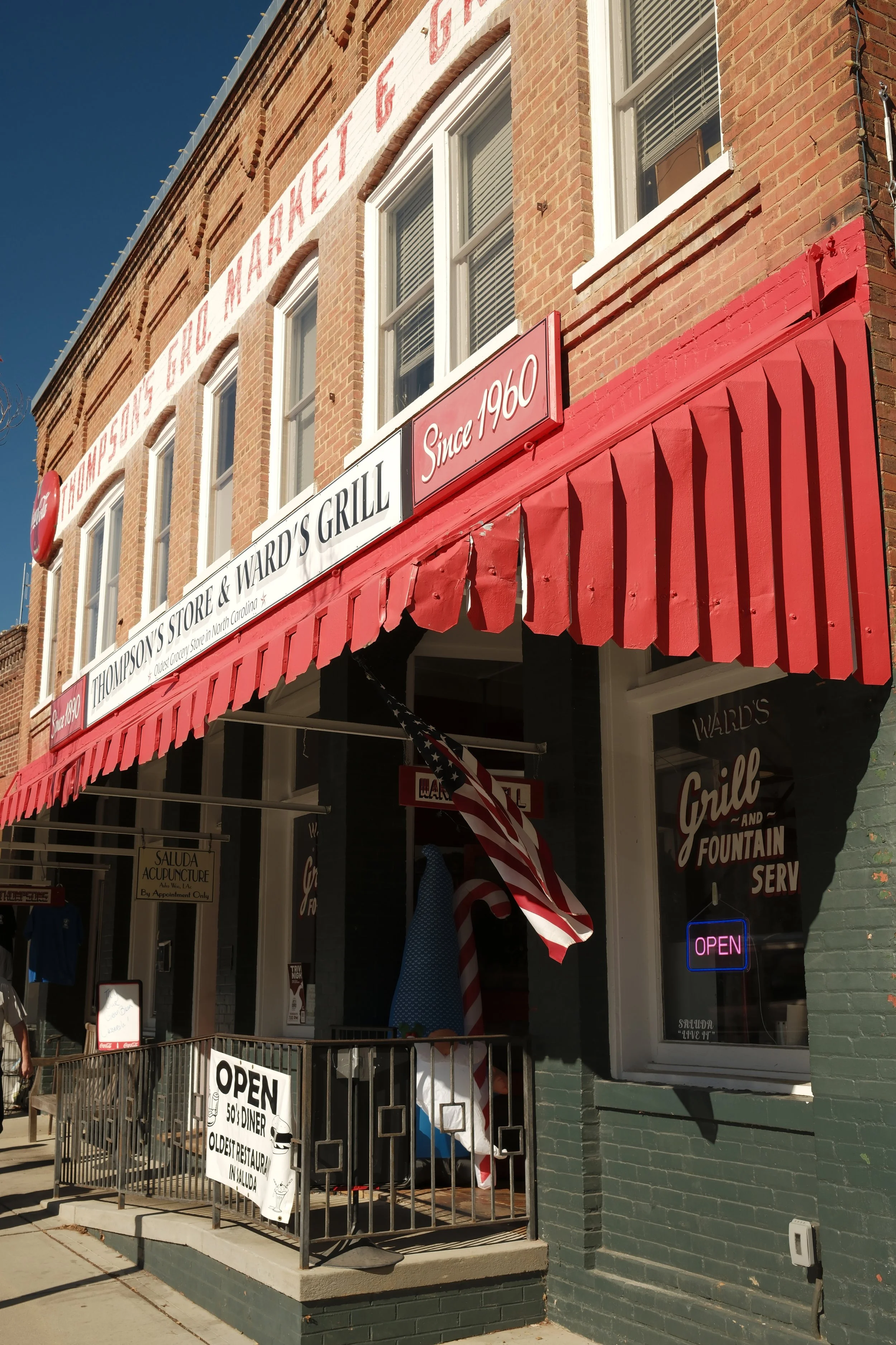 The exterior of Ward's Grill and Fountain, a restaurant established in 1960 with red brick walls and a red awning. The restaurant has multiple signs, including one that states 'Since 1960' and a neon 'Open' sign in the window. An American flag is dis