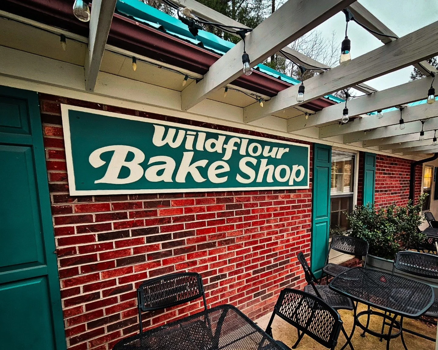 Outdoor seating area at Wildflour Bake Shop with black metal tables and chairs, brick wall, teal shutters, and a large teal sign with white text.