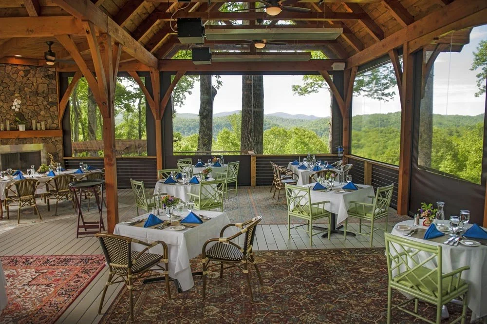 Outdoor dining area with round tables covered in white tablecloths, blue napkins, and glassware, set in a wooden pavilion with large windows overlooking a lush green forest and rolling hills.