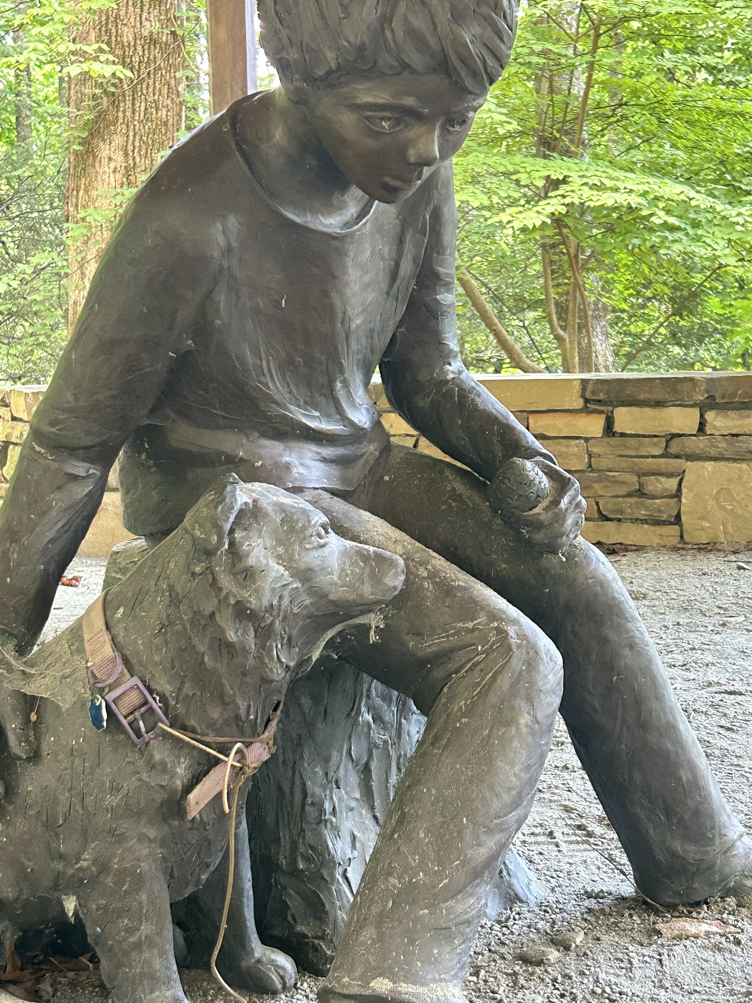 Bronze statue of a young girl sitting with her head slightly bowed, outdoors in a wooded area, with trees and green leaves in the background.