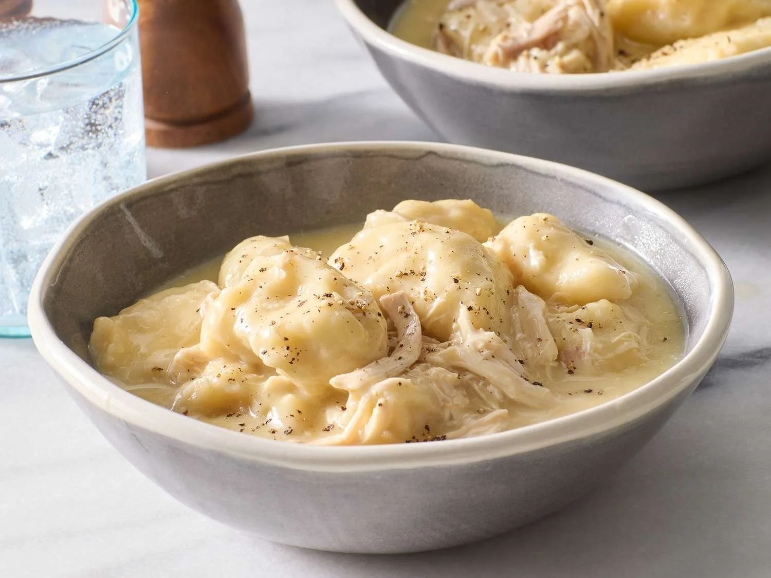 A bowl of mashed potatoes with gravy and black pepper, with a glass of water and a wooden salt shaker in the background.
