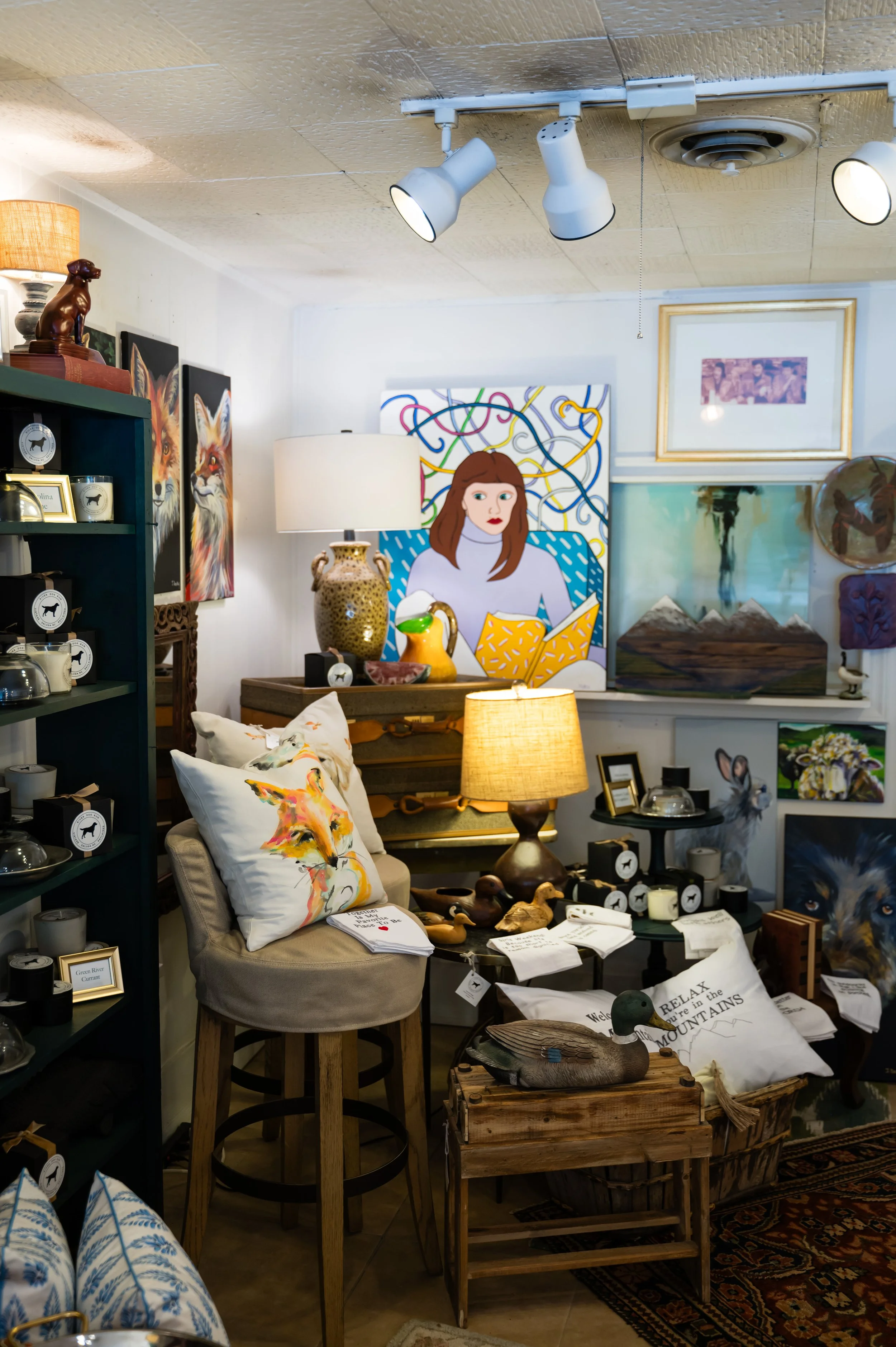 Interior of an art and home decor store with various paintings, decorative pillows, lamps, and vintage-style furniture, including a wooden chest and a small round table with decorative objects.