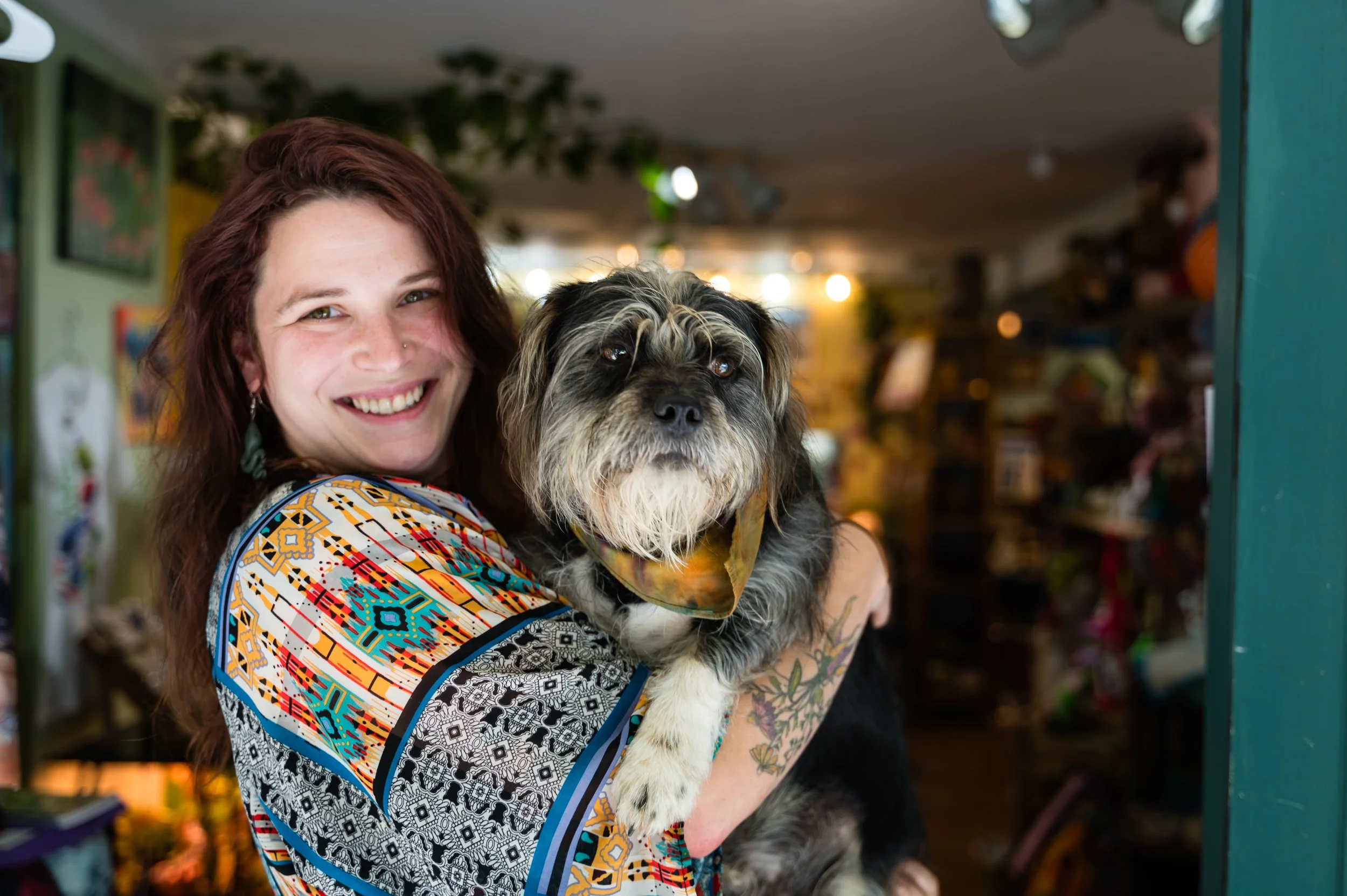 A woman with long reddish-brown hair smiling while holding a fluffy, black and white dog with a bandana around its neck inside a store.