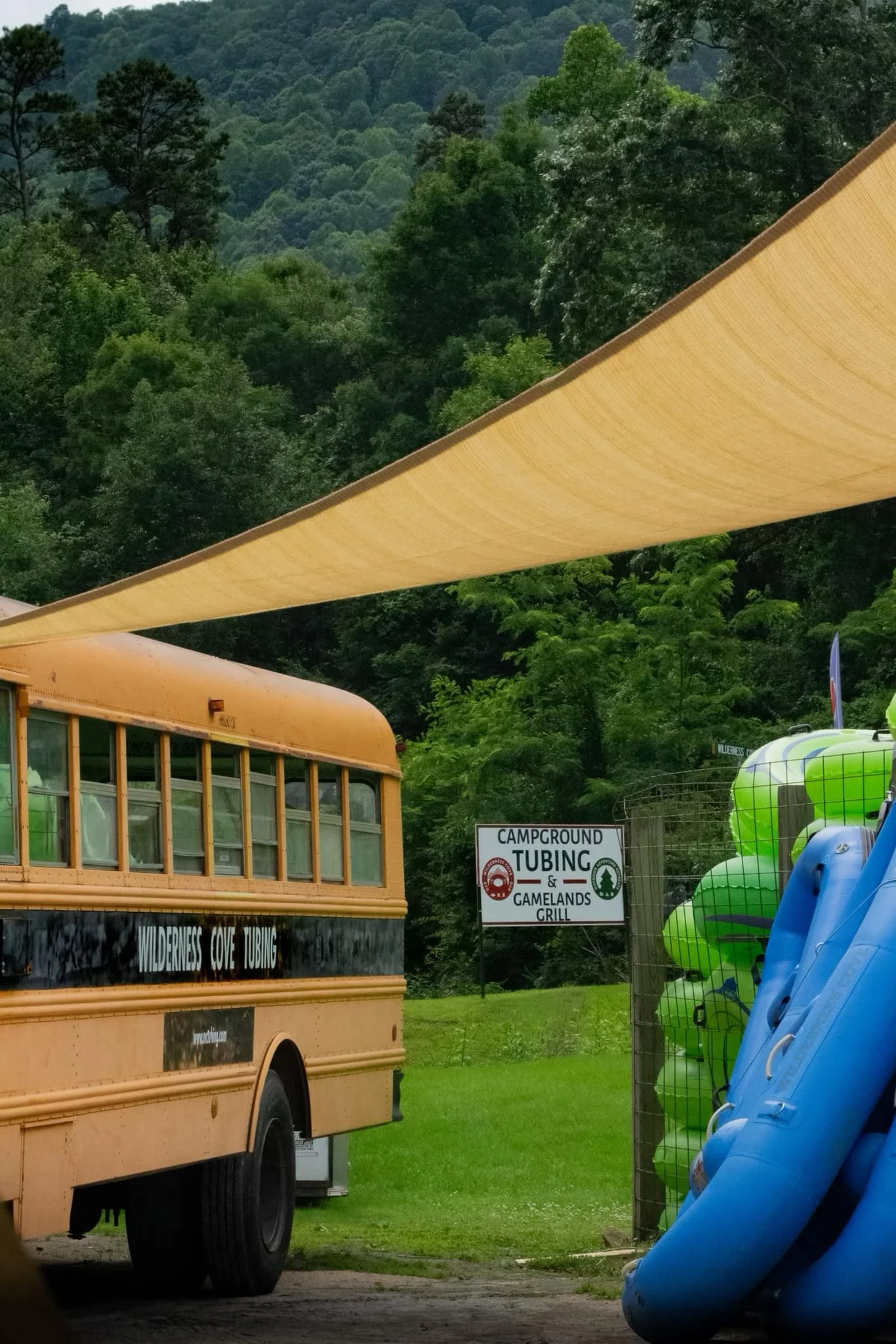 A yellow school bus parked next to a sign reading 'CAMPGROUND TUBING & GAME & GRILL' near a row of colorful kayaks in blue, green, and orange at a campground surrounded by lush green trees and hills.