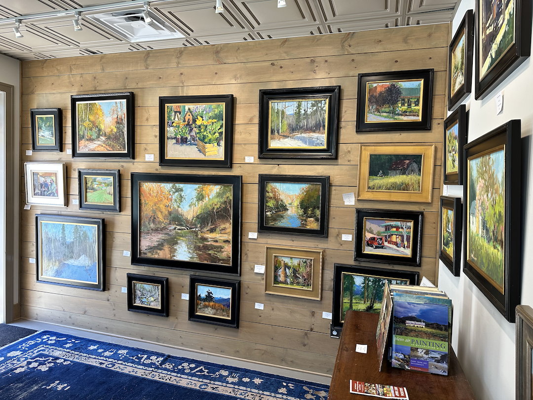 An art gallery display with various landscape paintings on a wooden wall and a few on an adjacent white wall, with a table below holding art brochures.