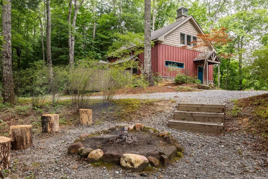 A rustic house in the forest with a gravel path, wooden steps, and a fire pit surrounded by rock and tree stumps.