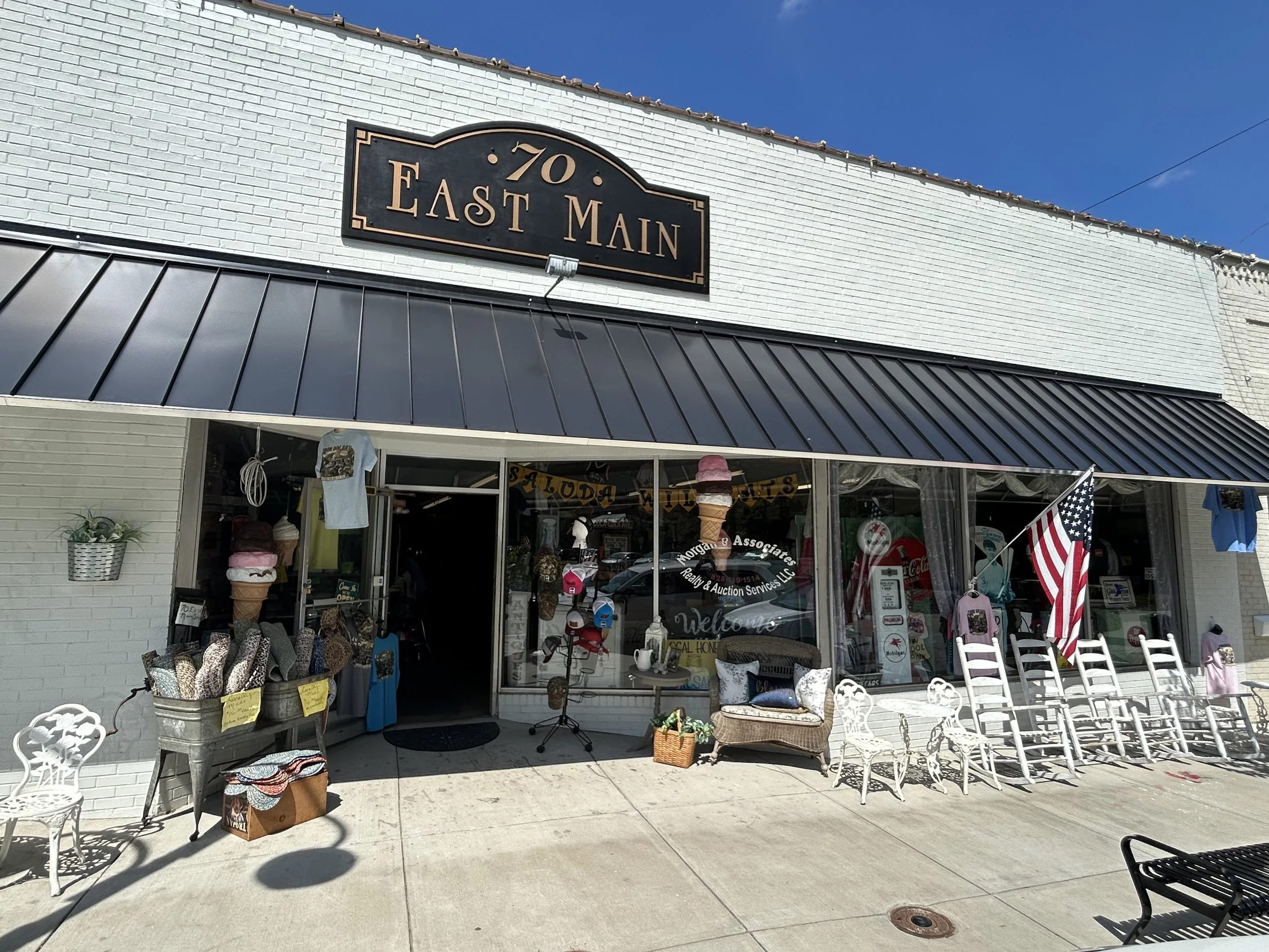 A storefront with a sign that reads '70 East Main' above the entrance. The shop displays various household items, like chairs, tables, and decorative objects, outside the store on the sidewalk. There is a patriotic American flag, and the store has la