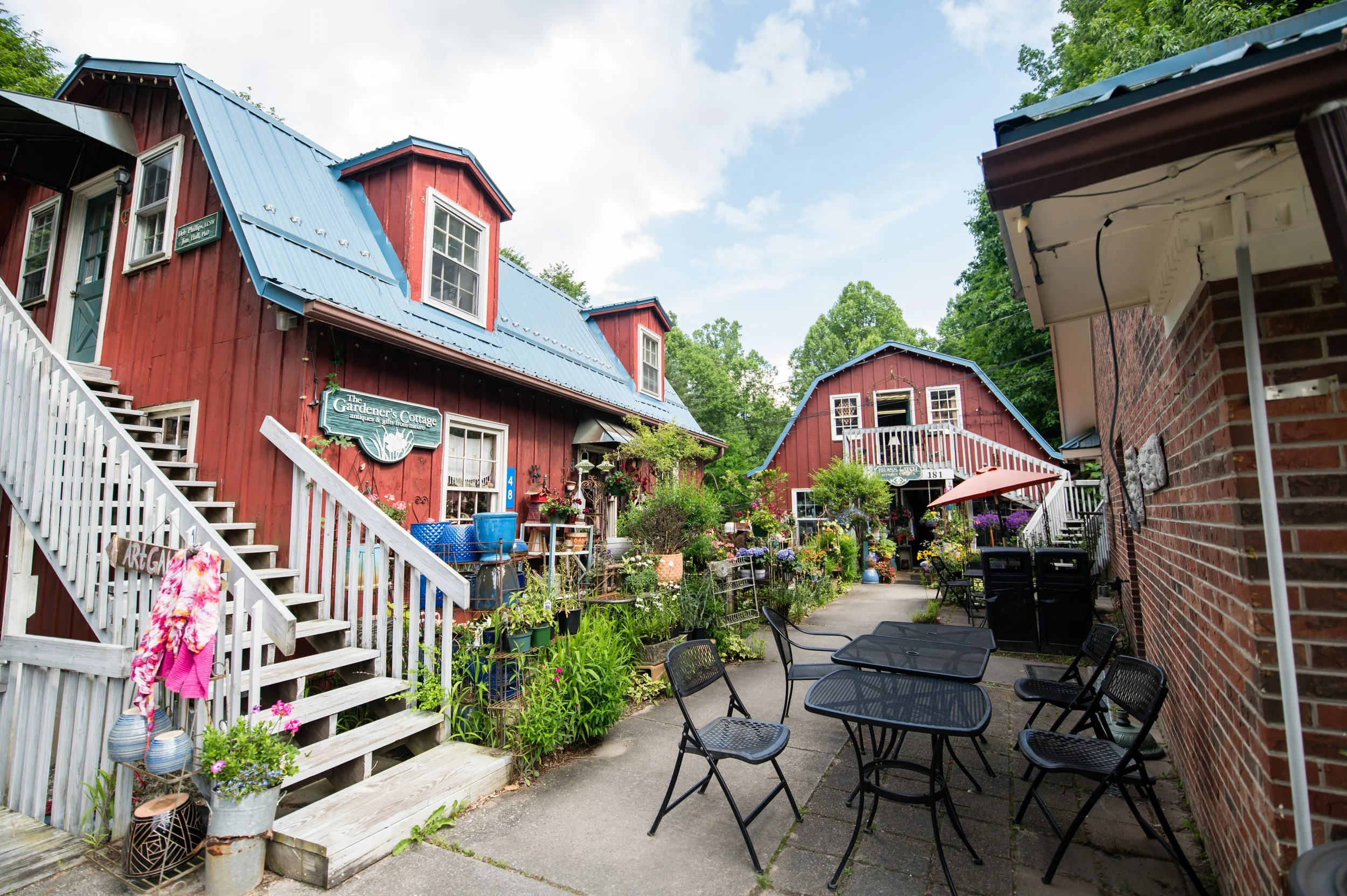 Outdoor patio with black metal tables and chairs, surrounded by plants and flowers, red buildings with blue roofs, and trees in the background under a partly cloudy sky.