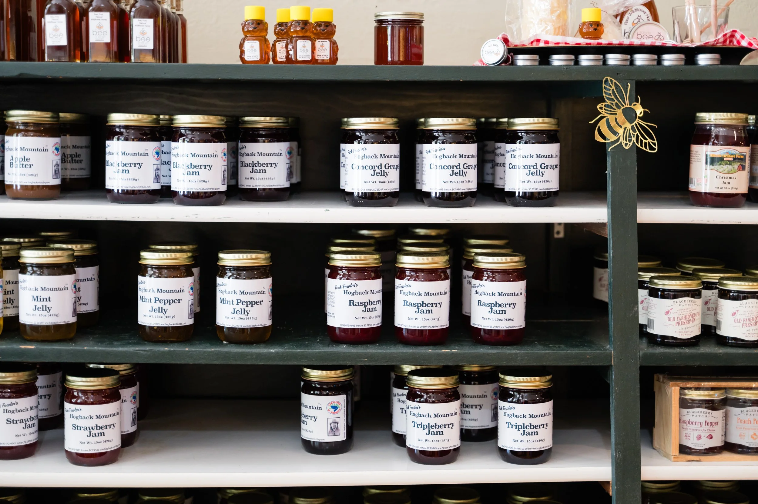 Shelves filled with jars of jam and preserves, including blackberry jam, blueberry jam, raspberry jam, and triple berry jam, with jars labeled and some topped with metal lids, in a store or market setting.