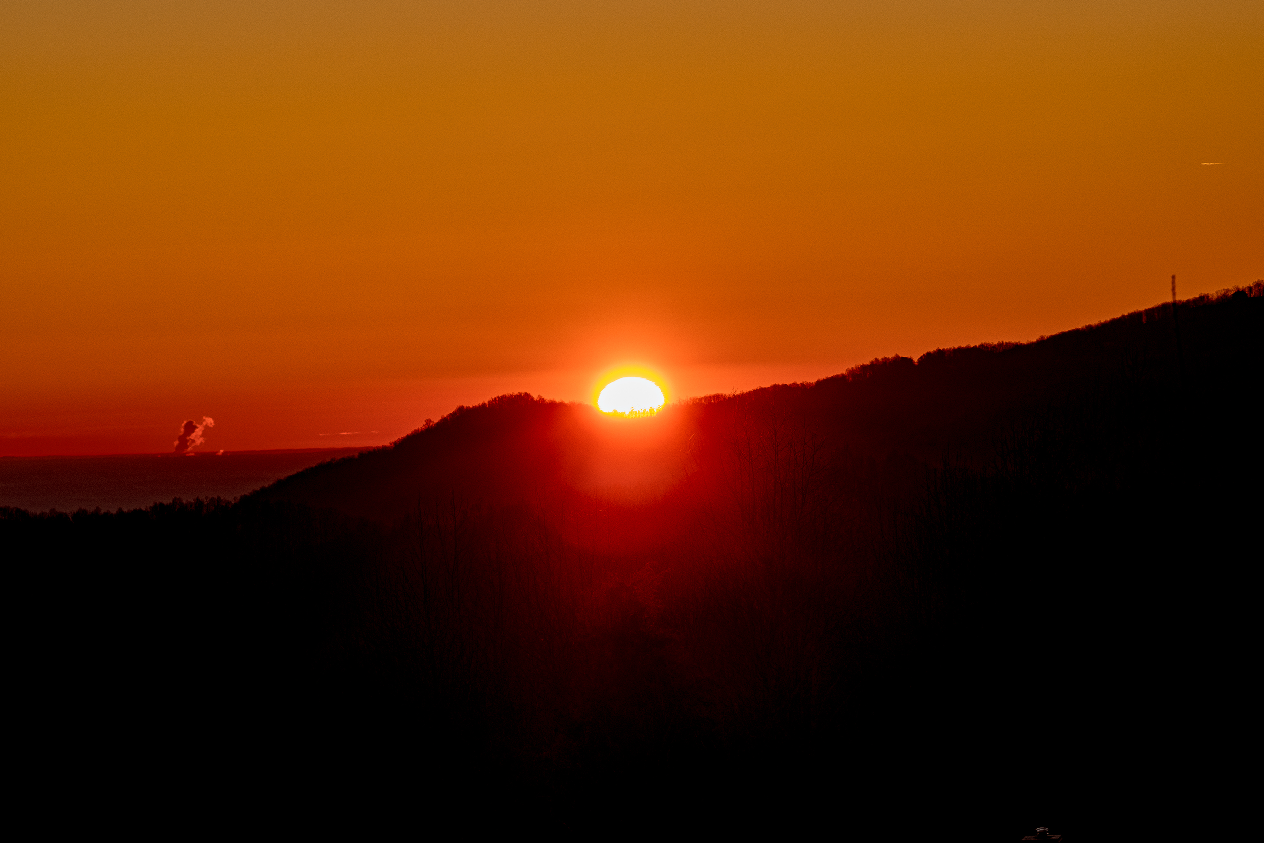 Sun setting behind a hillside with a clear orange sky and a few clouds in the distance.