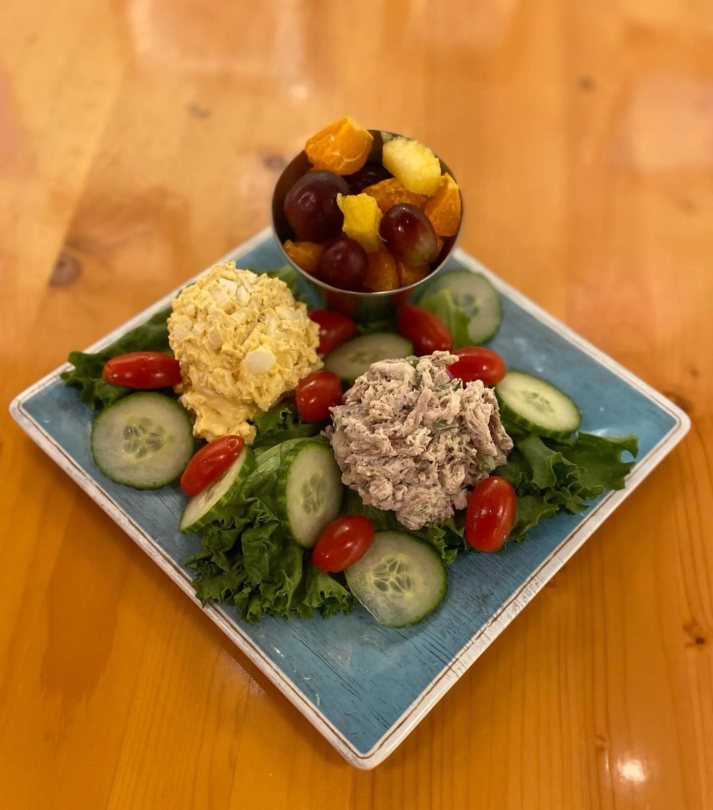A plate with leafy greens, sliced cucumbers, cherry tomatoes, and two scoops of tuna and chicken salad, garnished with grapes and dried fruit in a small cup.
