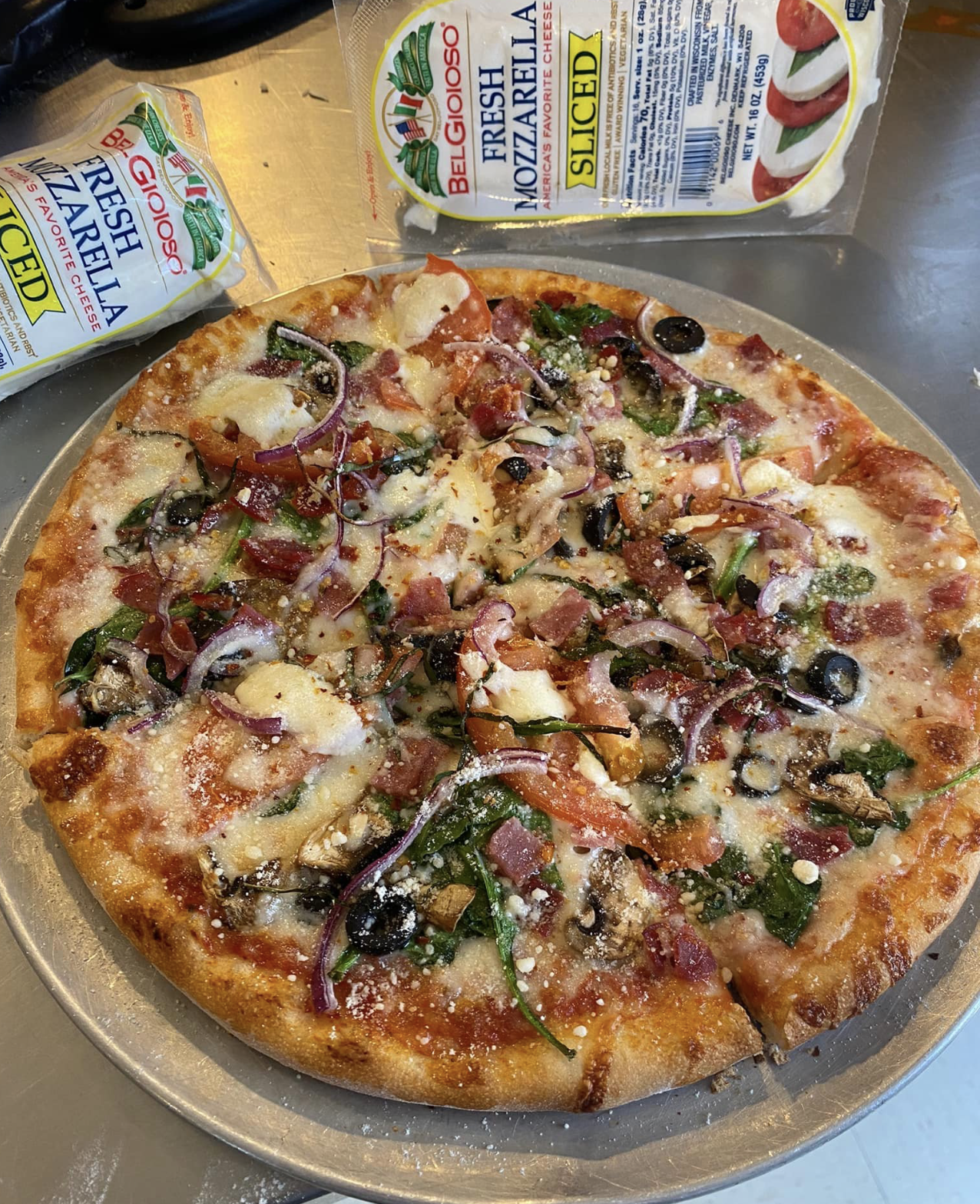 A pizza with cheese, black olives, onions, green peppers, and pepperoni on a silver tray, with packages of BelGioioso fresh mozzarella cheese in the background.