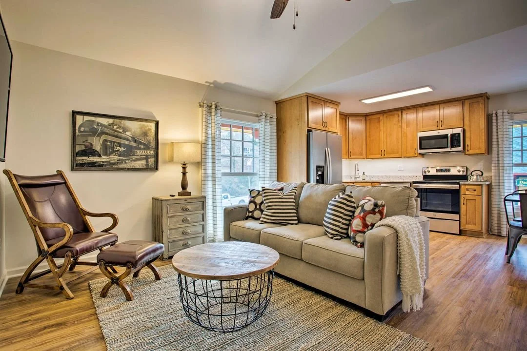 A cozy living room with a view into the kitchen, featuring a beige sofa with patterned pillows, a wooden coffee table with a metal wire base, a vintage armchair with a matching ottoman, a side table with a lamp, window with white curtains, and kitchen cabinets with stainless steel appliances.