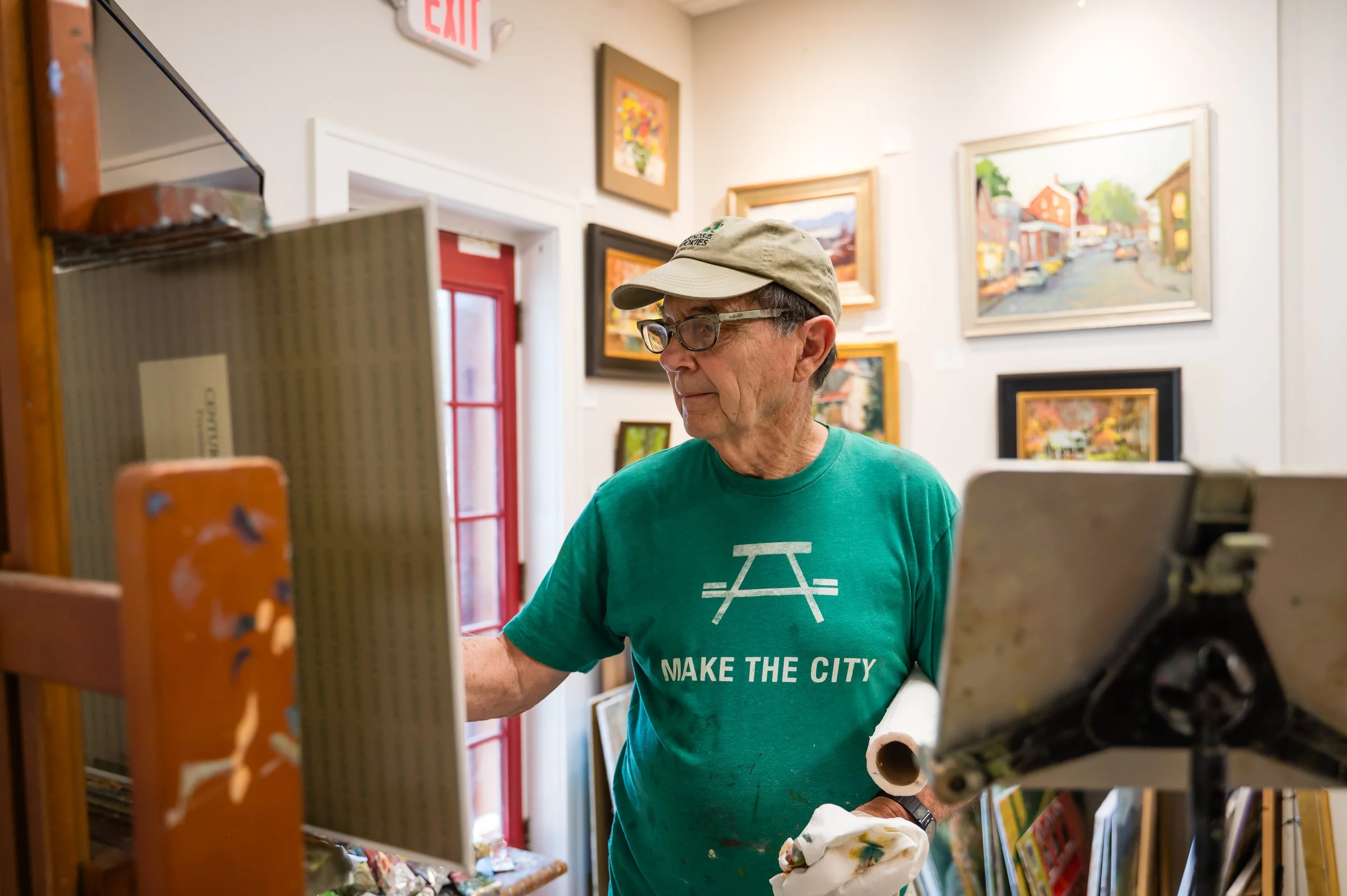 An elderly man with glasses wearing a green T-shirt with the words 'MAKE THE CITY' and a graphic of a bridge, browsing artwork in an art gallery.