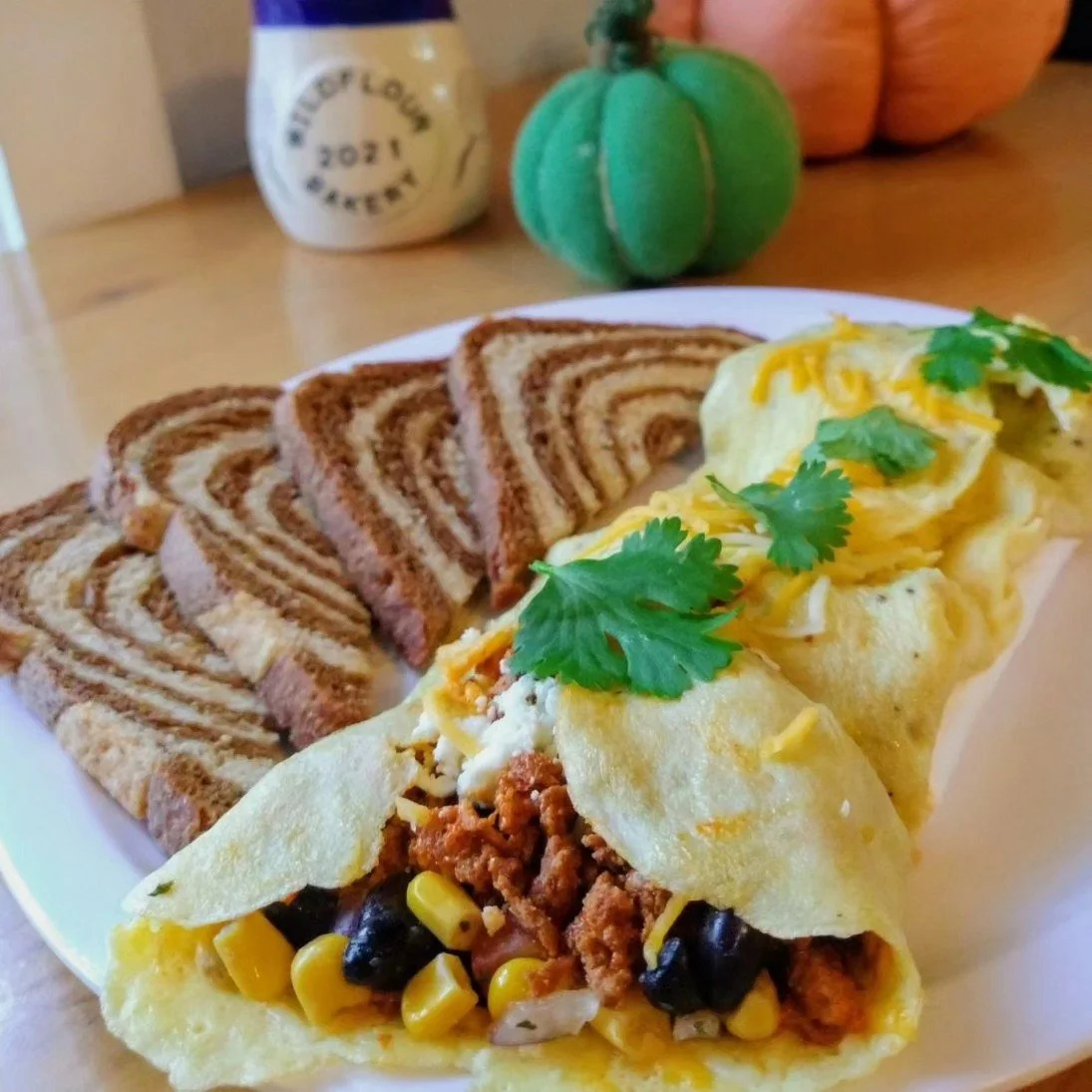 A plate with two breakfast tacos filled with seasoned meat, corn, black beans, cheese, and garnished with cilantro, alongside slices of cinnamon swirl bread. In the background, there is a green plush pumpkin, a jar labeled "Top of the Hour 2021," and