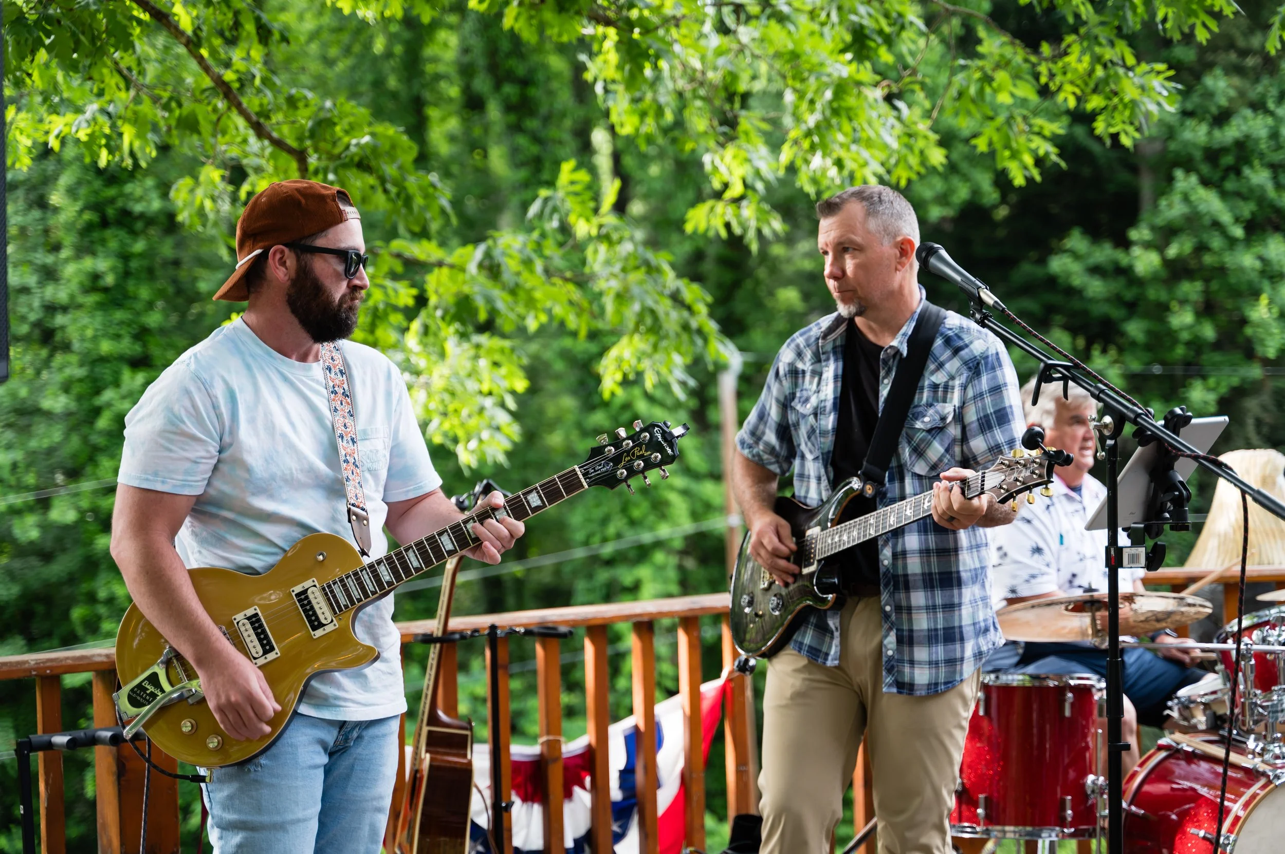 Two men playing guitars in an outdoor setting with green trees, one wearing a white shirt with sunglasses and a backward cap, the other in a plaid shirt with short gray hair, with a drum set in the background.