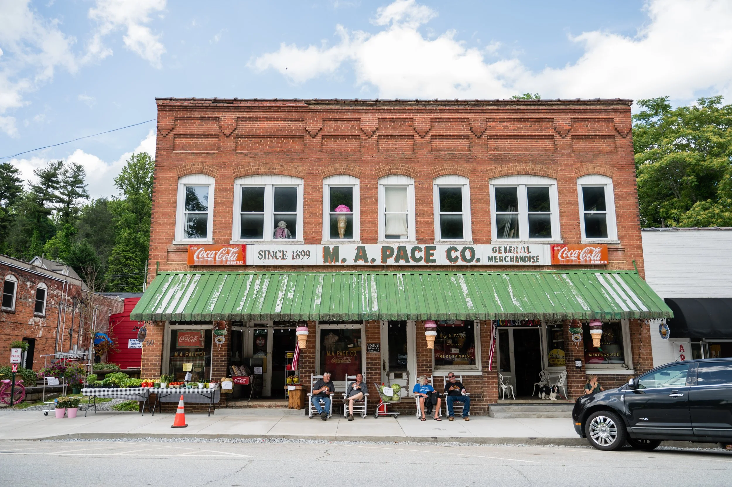 A two-story brick building with a green awning and a storefront selling merchandise and food. There are four people sitting on chairs in front of the building, and a few small tables with flowers and watermelon dishes. The upper floor has five windows with decor, and there are signs indicating the store is M. A. Pace Co., established in 1899, selling general merchandise. There are also Coca-Cola signs on the building.