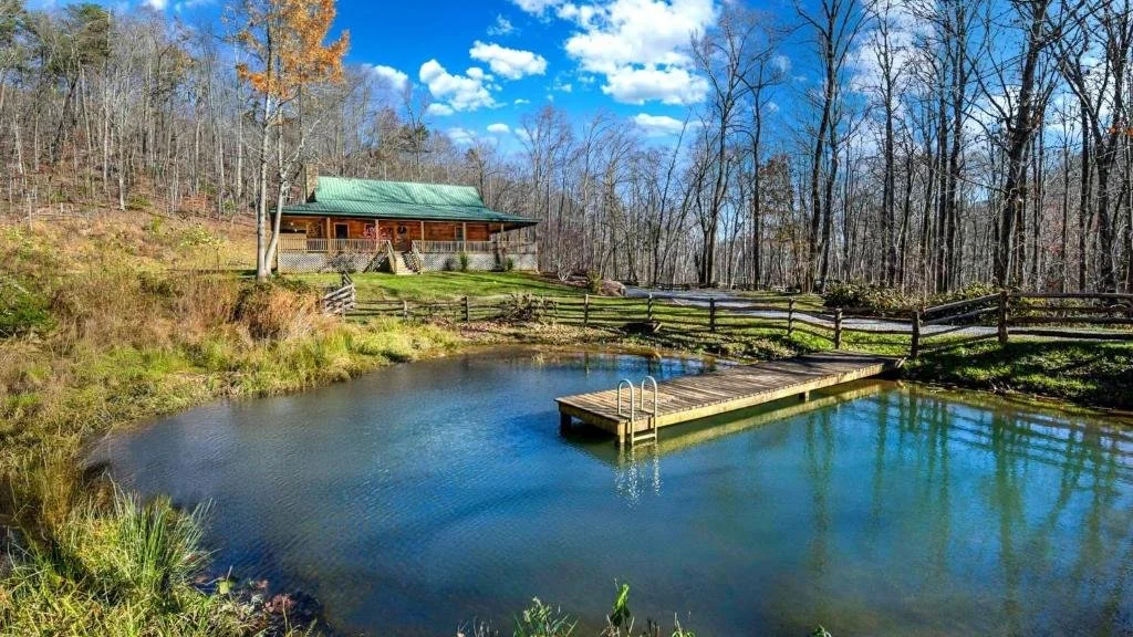 A cabin with a green roof sits on a hill beside a small pond with a dock. The scene is surrounded by leafless trees under a blue sky with scattered clouds.