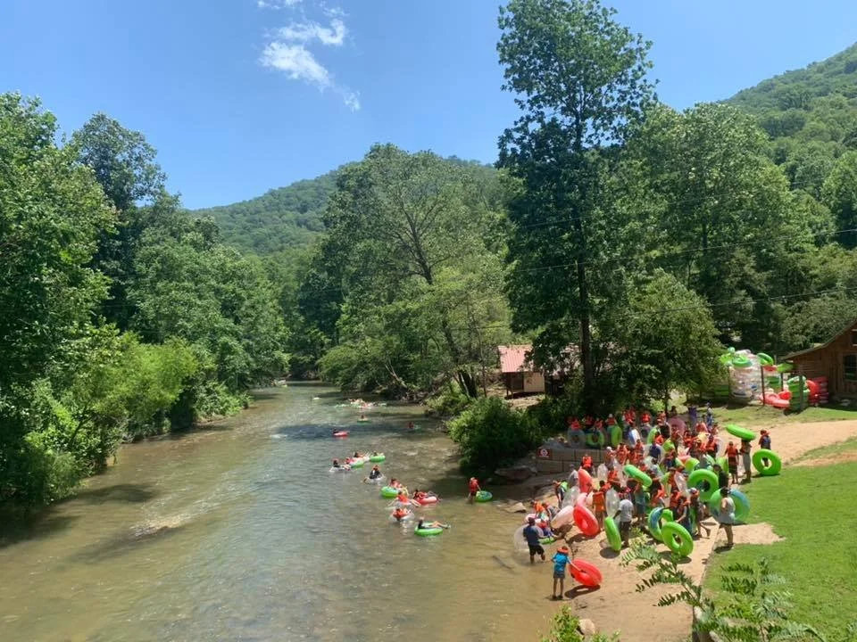 People swimming and playing with inflatable tubes in a river beside a forested area with a small cabin and large inflatable water toys on the grass.