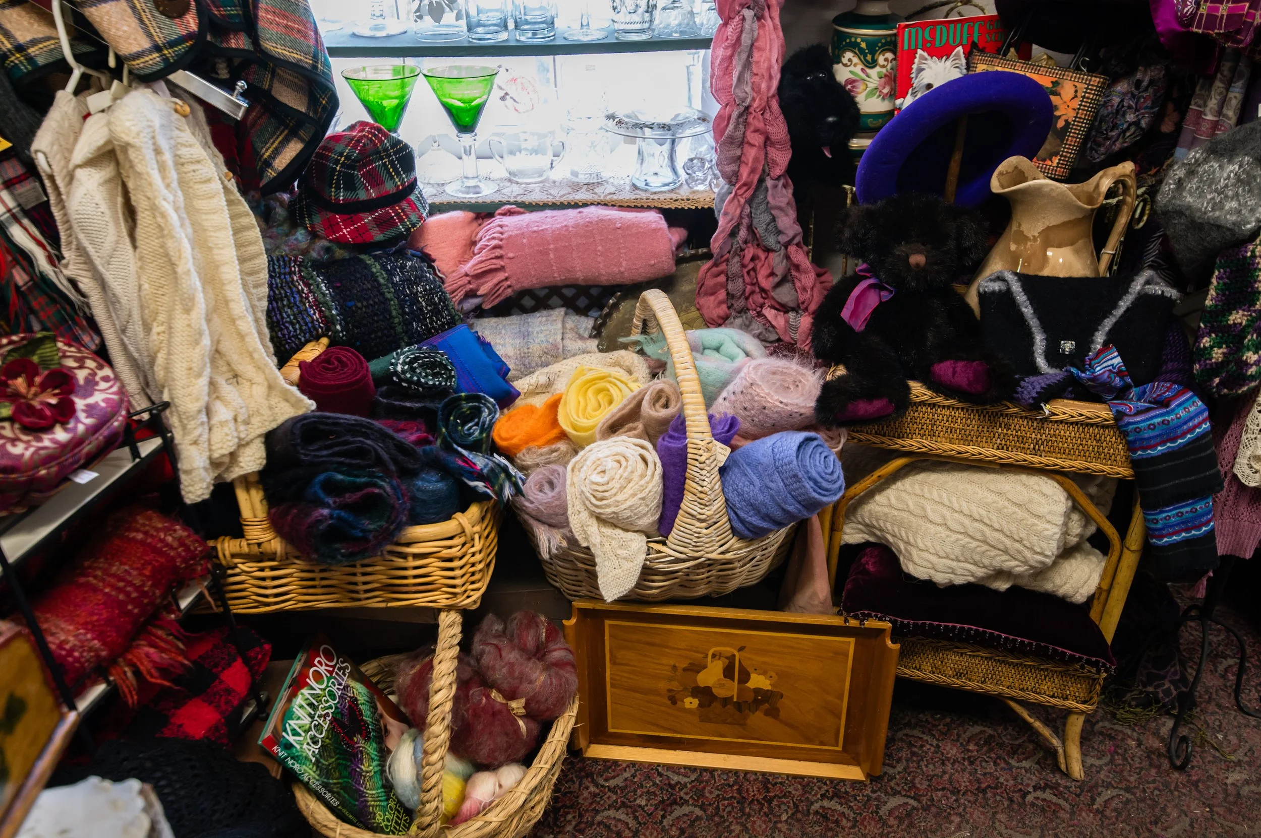 A display of various small retail items including yarn, scarves, and hats in a thrift or craft store, with a black dog wearing a pink collar among the products.