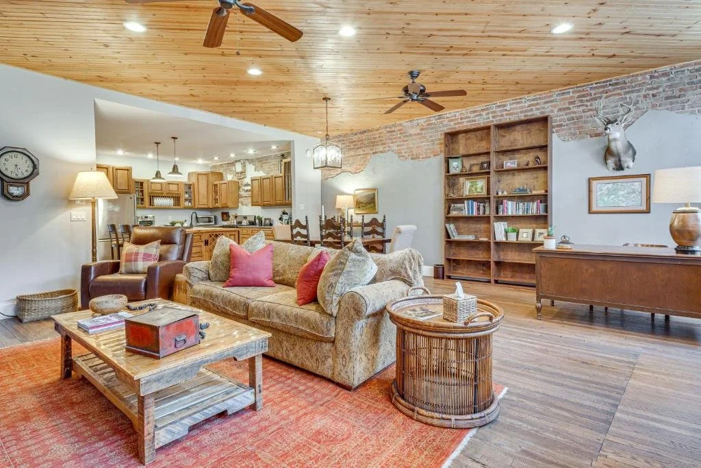 Living room with wooden ceiling, brick wall, and wood floor, furnished with a floral sofa, leather armchair, wooden coffee table, bookshelf, and decorative items.