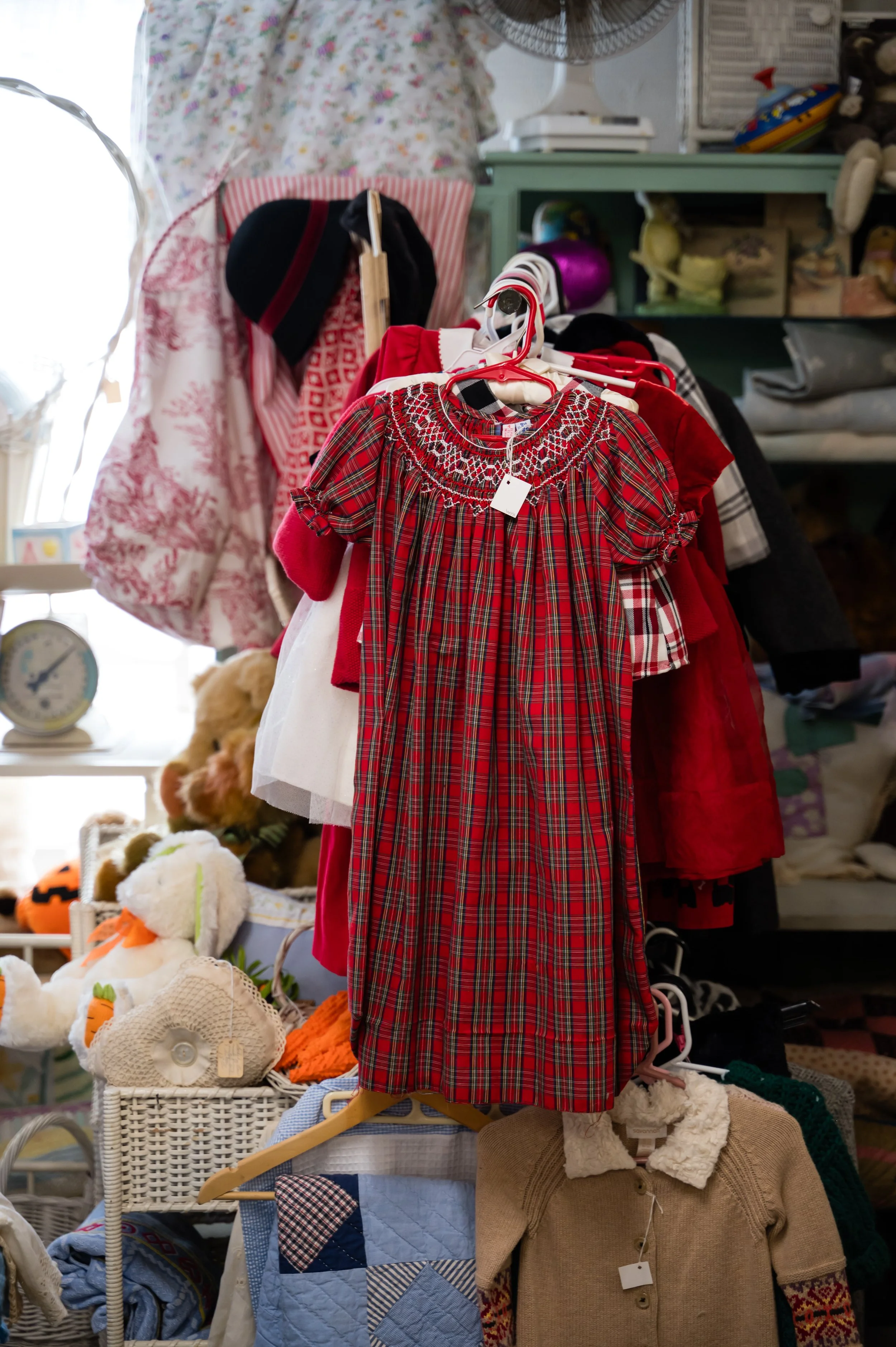 Clothing display with a red plaid dress on a hanger, surrounded by various children's garments and toys in a thrift or secondhand store.