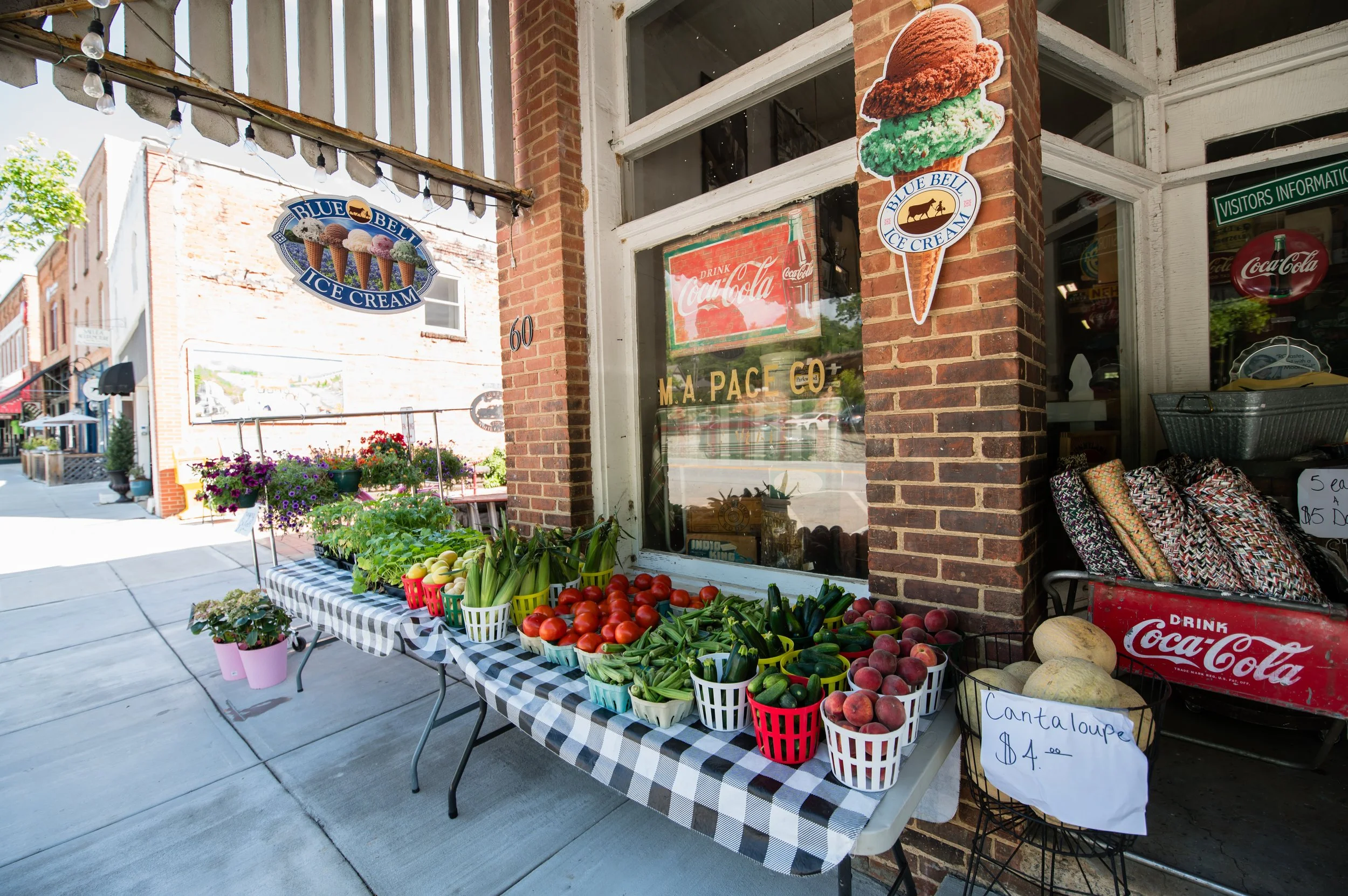 Street view of a fruit and vegetable stand outside a brick storefront. The stand has a checkered tablecloth, and displays tomatoes, peaches, green beans, zucchini, and other produce in baskets. Signage includes ice cream advertising, a Coca-Cola sign