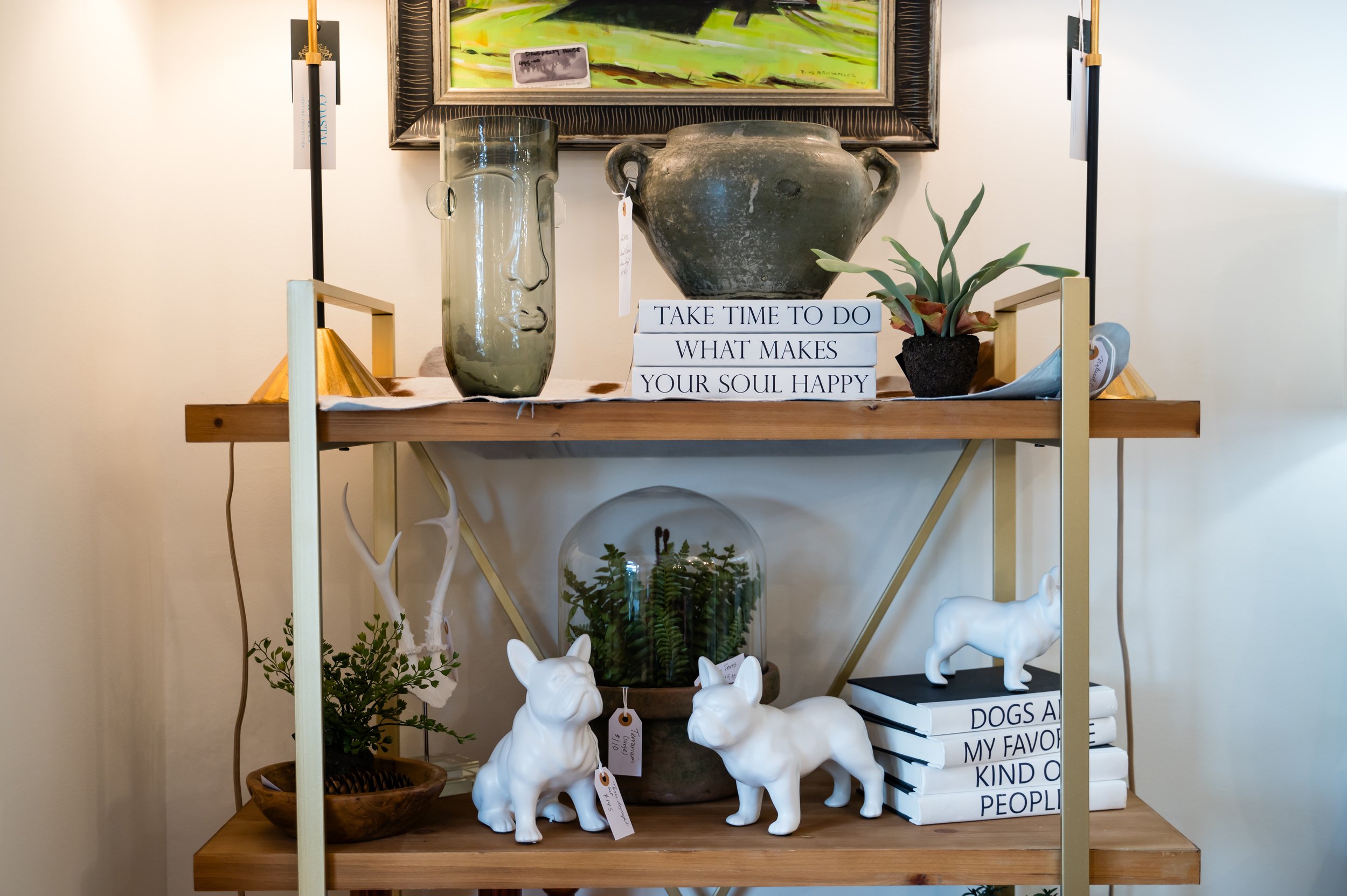 Decorative shelf with a large gray urn, a tall glass vase with face design, potted plant, books with motivational titles, and white ceramic animal figurines, all against light-colored wall with framed artwork.