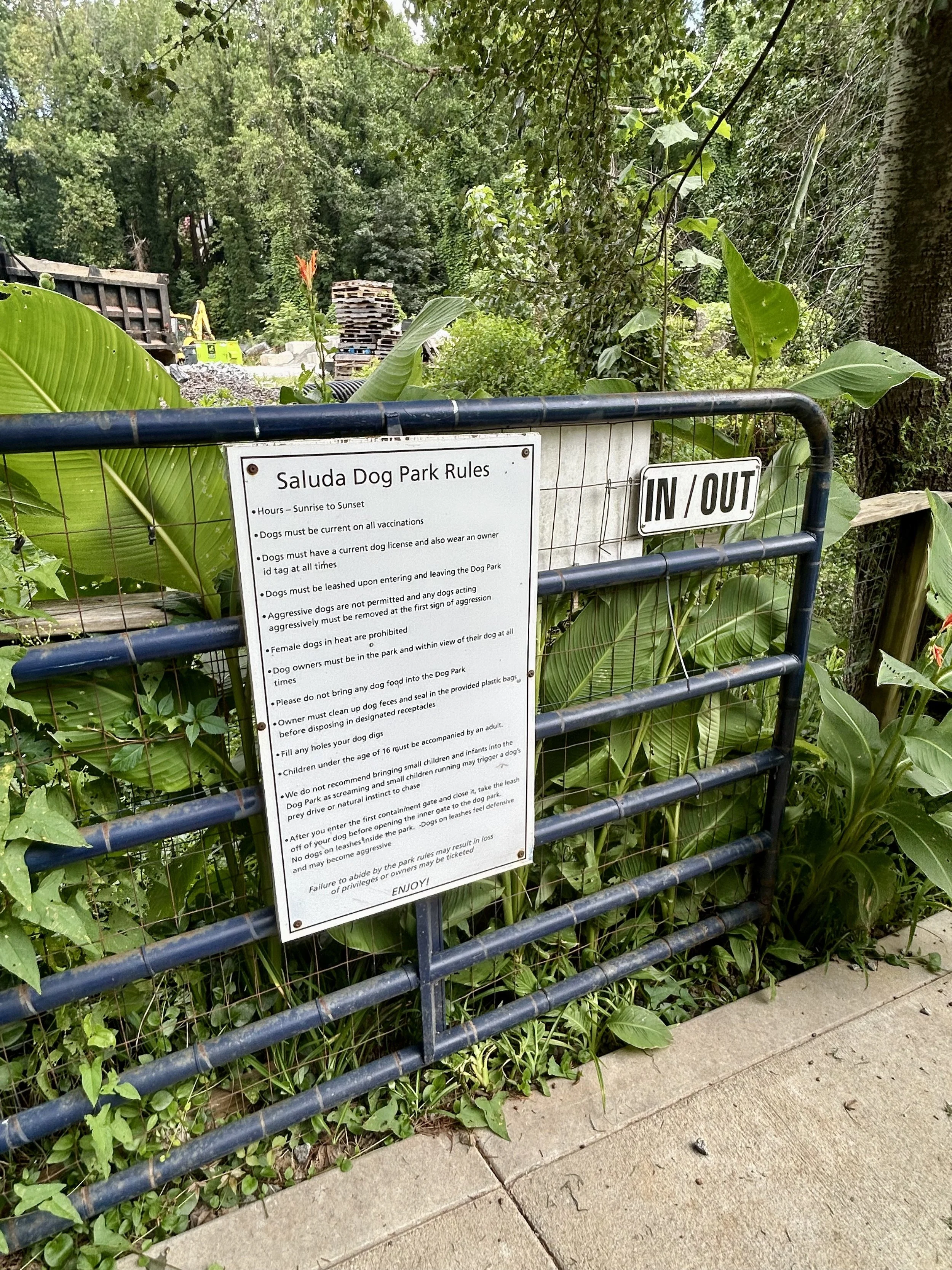 A sign at Saluda Dog Park displaying rules for dog owners, attached to a black metal fence with lush green plants and trees in the background.
