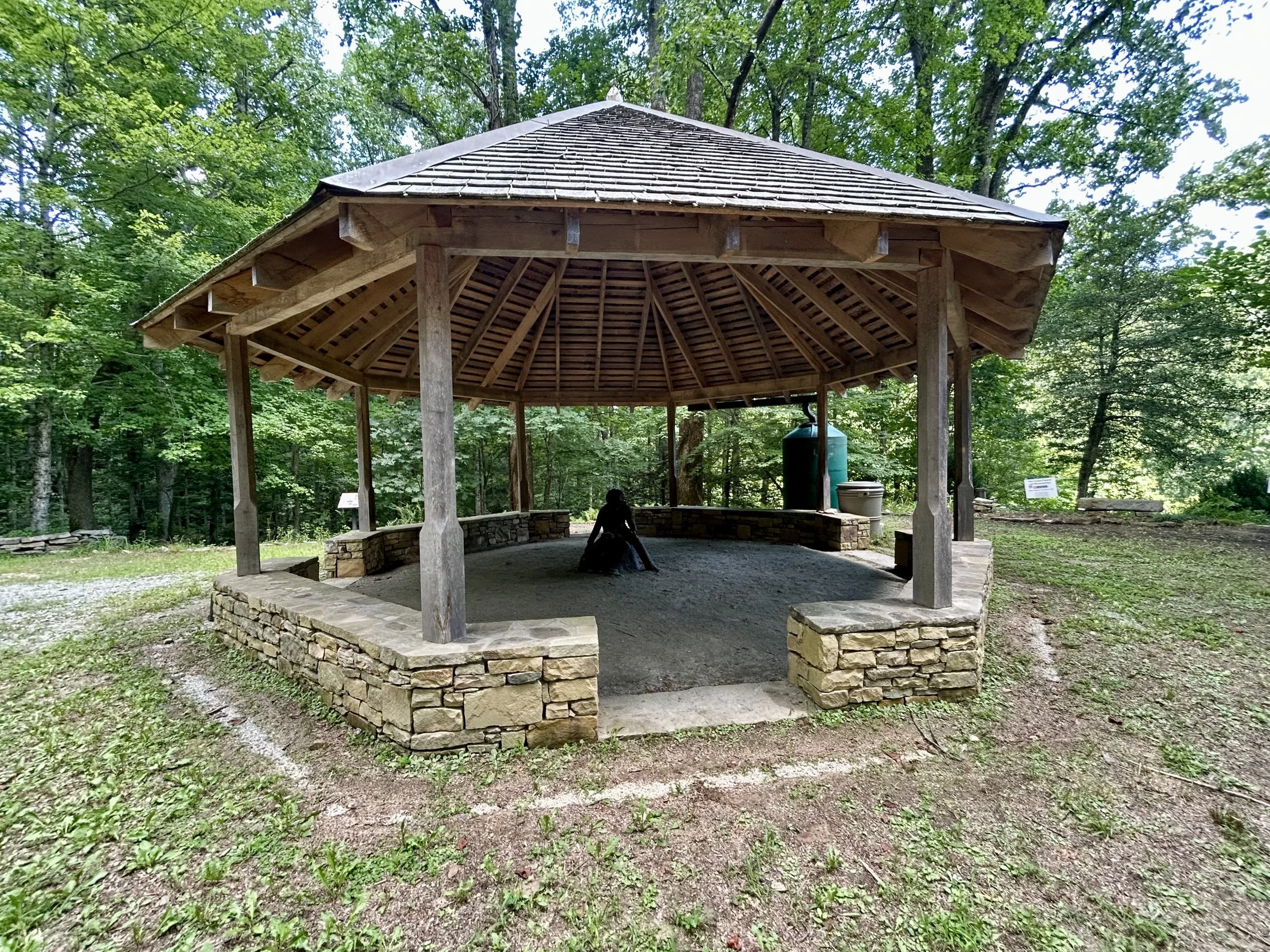 A wooden shelter with a shingled roof surrounded by green trees in a forest, with a person sitting underneath.