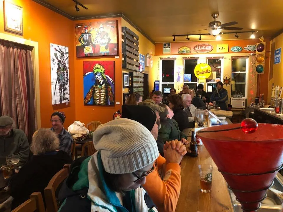 A lively bar with people sitting and enjoying drinks. The walls are decorated with colorful artwork and signs, and there is a bar counter with glasses and bottles. The atmosphere appears cheerful and social.