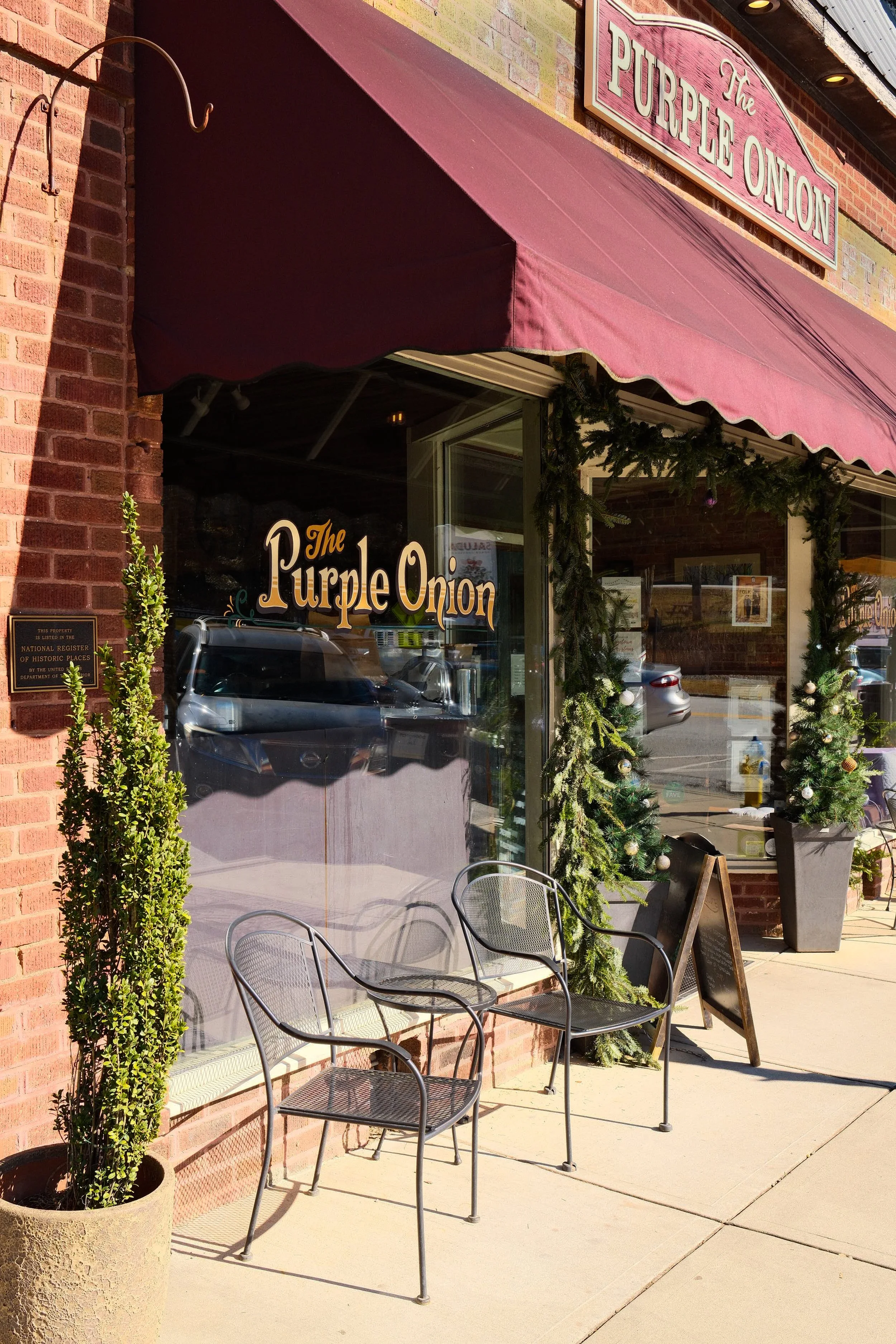 Exterior of a restaurant named 'The Purple Onion' with a maroon awning, decorated with Christmas greenery. There are metal chairs and a large potted plant outside.