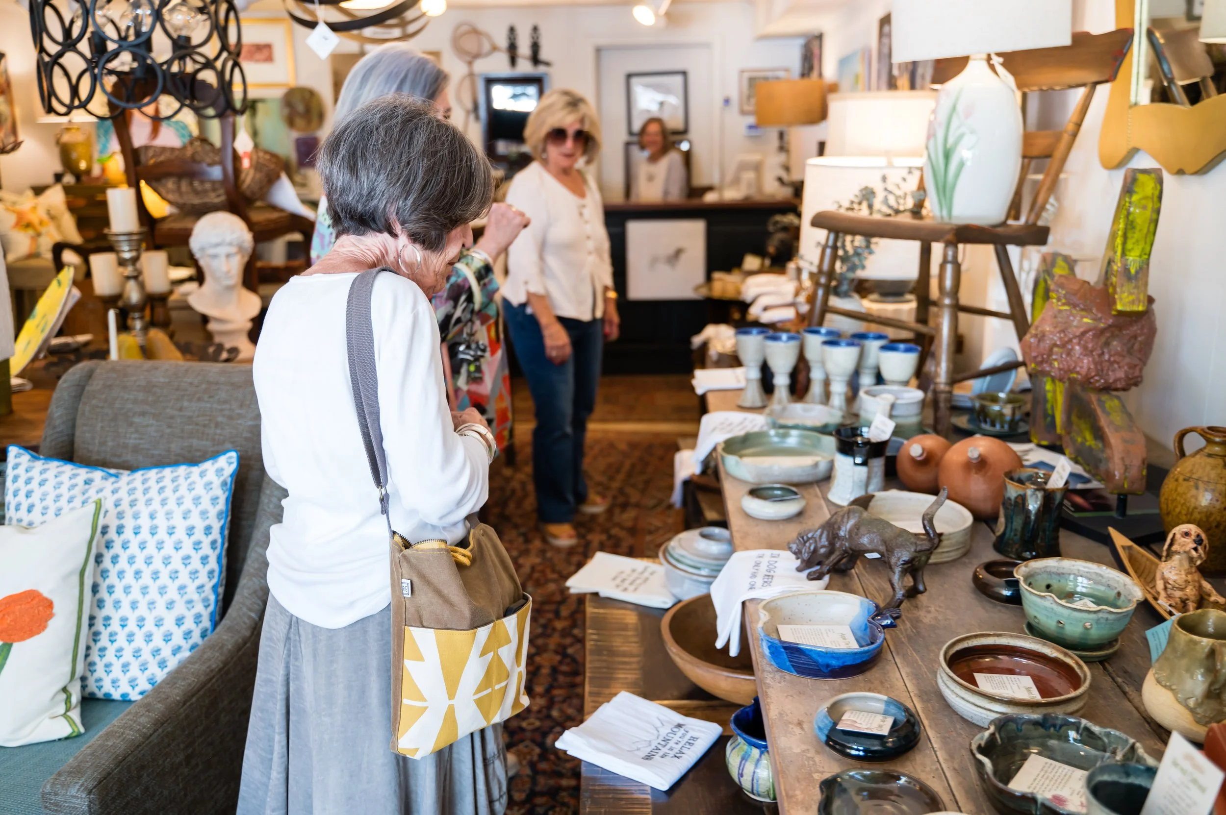 People browsing pottery and art pieces at an indoor craft fair or store, with shelves and tables displaying ceramics, sculptures, and artworks.