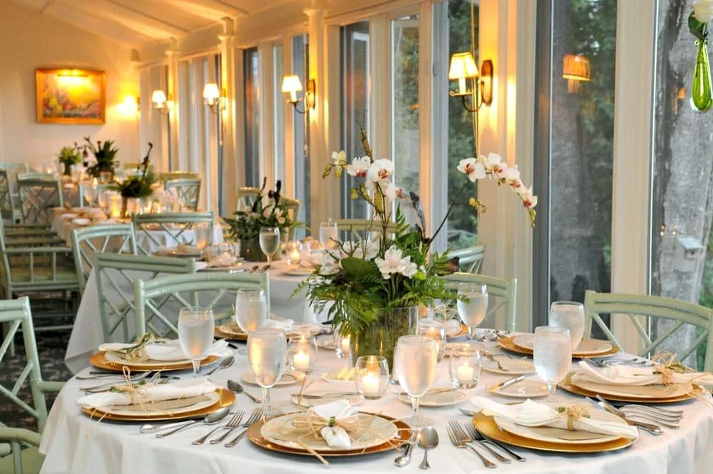 A beautifully decorated indoor dining area with multiple round tables set with white tablecloths, gold chargers, white napkins tied with twine, glassware, and cutlery. Each table has a centerpiece with white orchids and greenery, and small candles or tea lights. Large windows along the wall provide natural light and a view outside. The room features wall sconces and a framed picture on the wall.