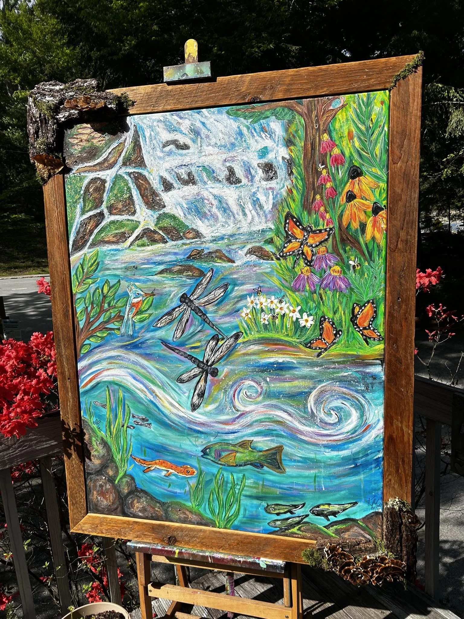 Colorful painting of a waterfall, flowers, dragonflies, butterflies, and fish outdoors on an easel framed by wood and surrounding plants.